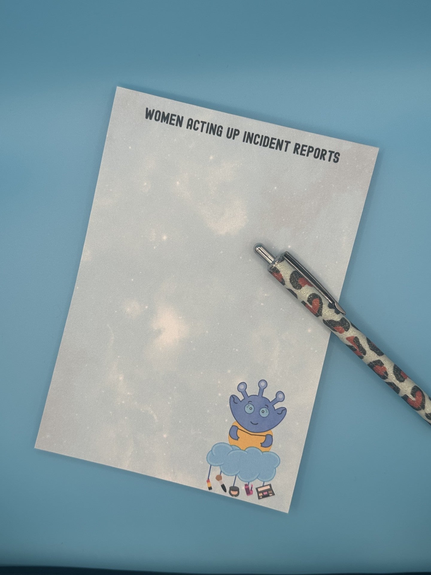 Notepad with character Cosmo Menace that looks like a blue alien floating on a cloud with makeup hanging from it. The notepad is a light blue sparkle background and has a heading on notepad that states "women acting up incident reports". The notepad is sitting on top of a blue background and a pen.