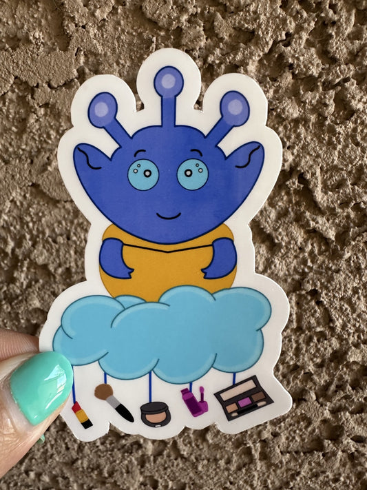 Blue alien sticker with a cloud and various makeup items hanging from the cloud. The background is on a textured surface. 