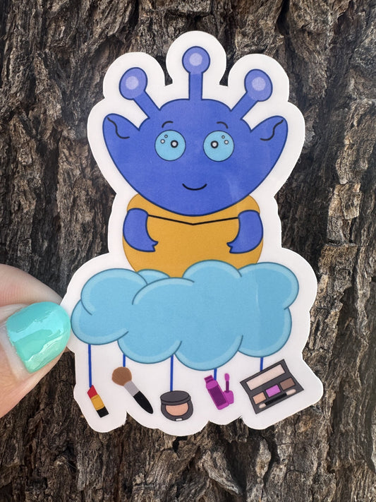 Blue alien sticker with a cloud and various makeup items hanging from the cloud. The background is a tree trunk.