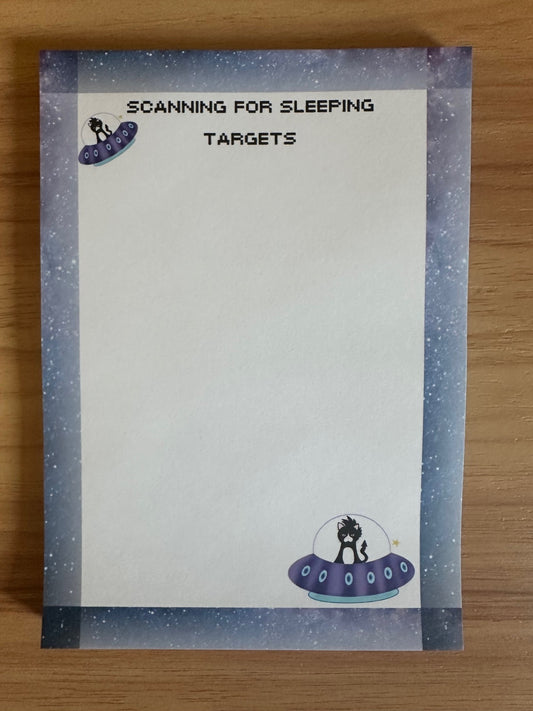 Notepad with fictional character that is a cat in a spaceship with headline on notepad that states "scanning for sleeping targets" sitting on a wooden surface. 