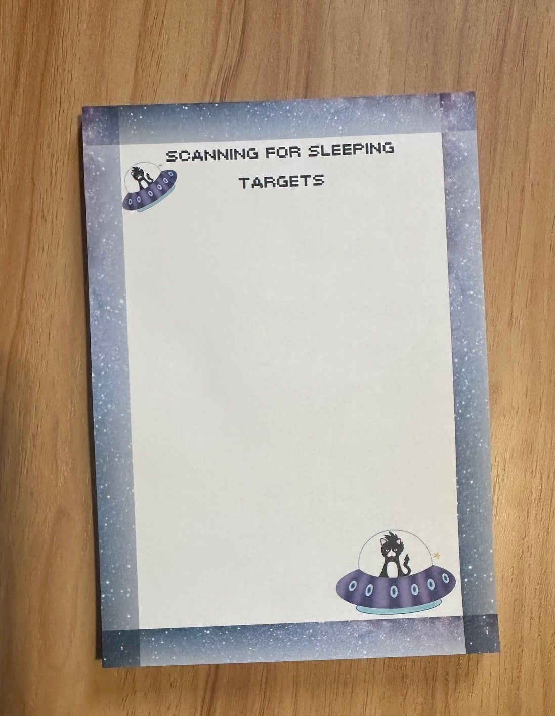 Notepad with fictional character that is a cat in a spaceship with headline on notepad that states "scanning for sleeping targets" sitting on a wooden surface. 