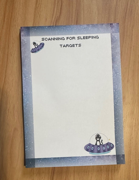Notepad with fictional character that is a cat in a spaceship with headline on notepad that states "scanning for sleeping targets" sitting on a wooden surface. 