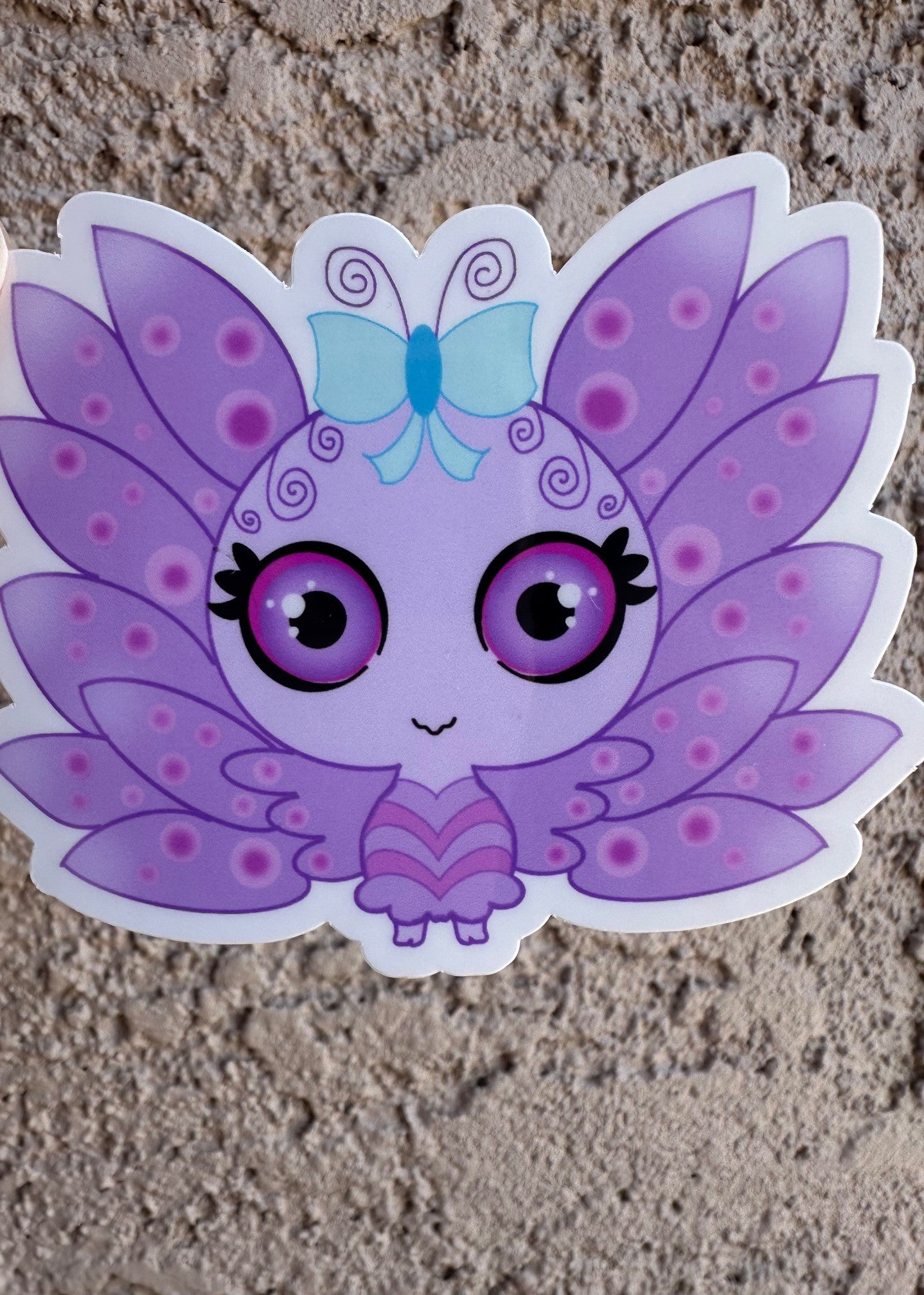 A vinyl sticker cartoon butterfly character with purple wings and a blue bow. The character has purple and black eyes with light purple circle patterns on the wings. The background is a textured brown background.