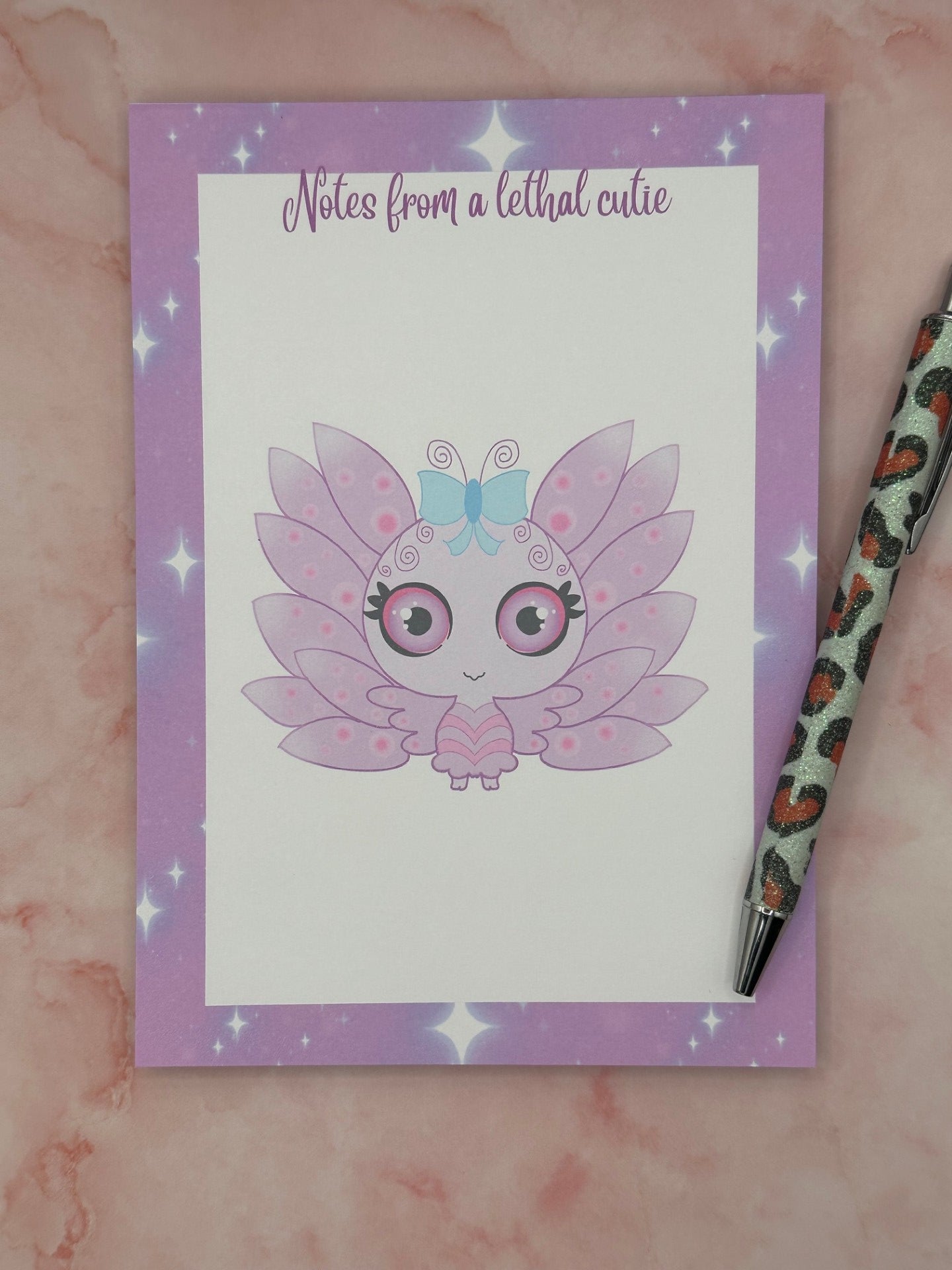 Notepad that has purple and sparkle edges around all four corners. The notepad heading states " Notes from a lethal cutie". The center of the notepad has a fictional character that is purple and pink with wings and a blue bow. The character has big black, purple and pink eyes. The notepad is sitting on top of a pink background with a pen. 
