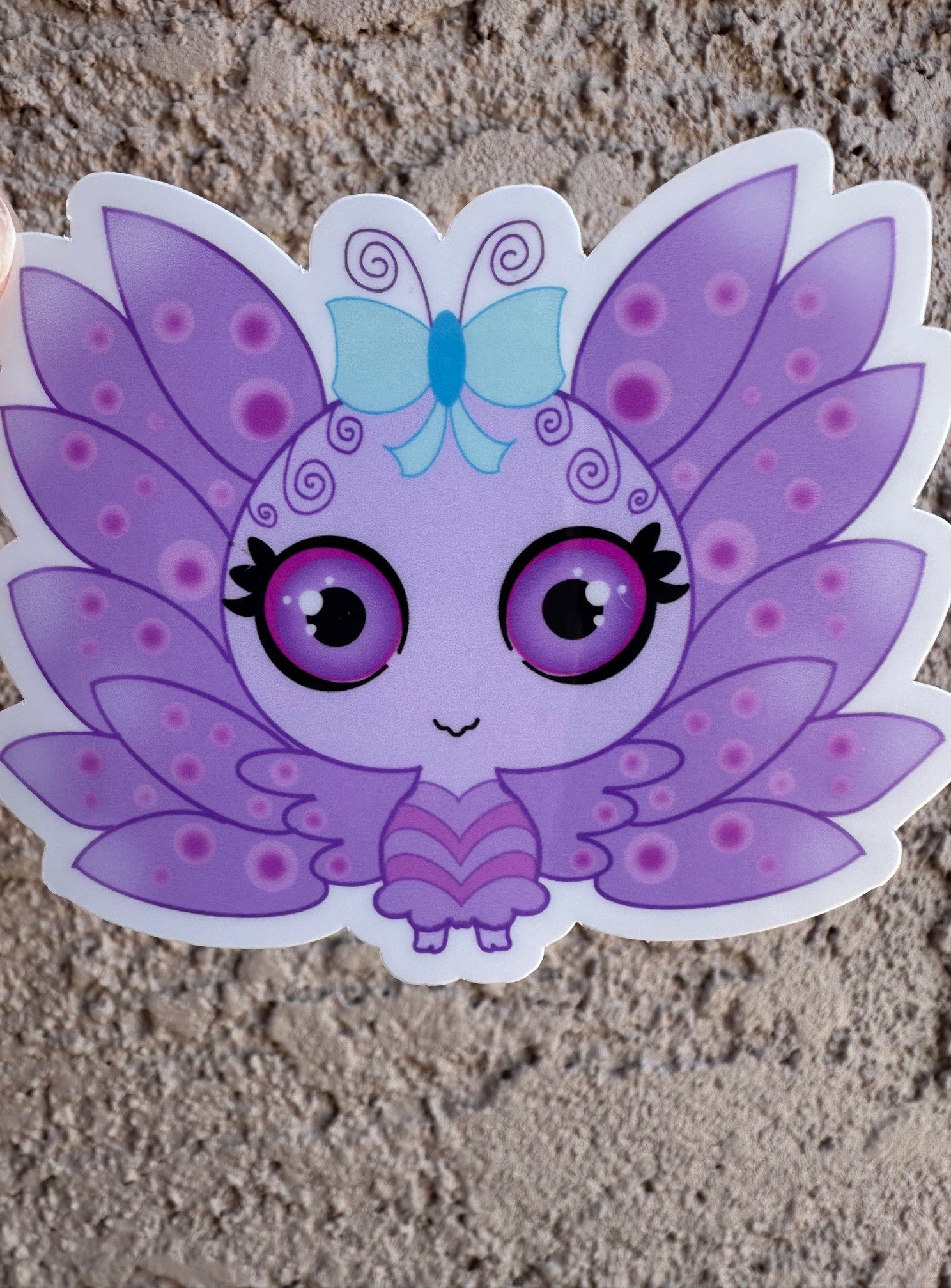 A vinyl sticker cartoon butterfly character with purple wings and a blue bow. The character has purple and black eyes with light purple circle patterns on the wings. The background is a textured brown background.