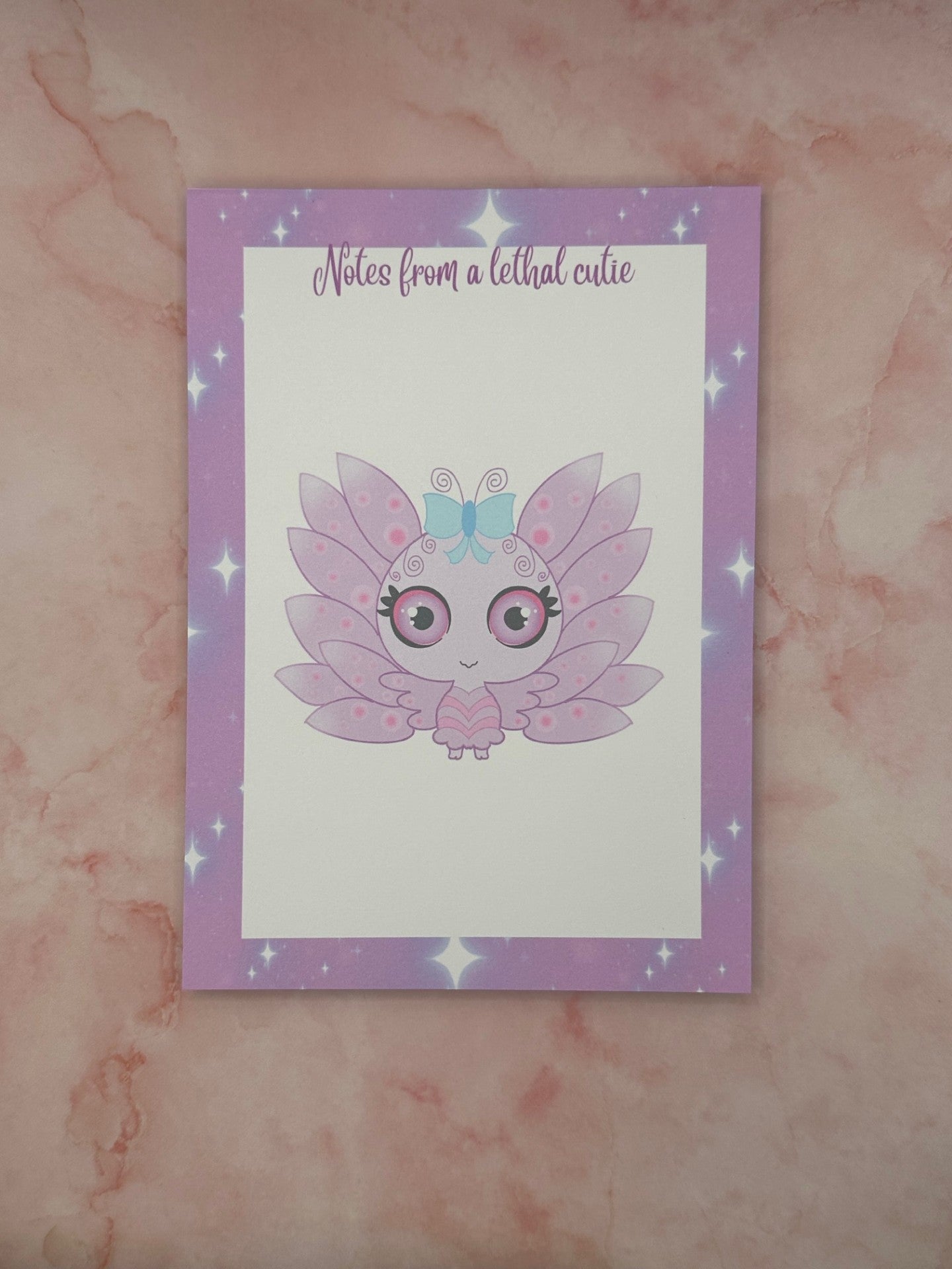 Notepad that has purple and sparkle edges around all four corners. The notepad heading states " Notes from a lethal cutie". The center of the notepad has a fictional character that is purple and pink with wings and a blue bow. The character has big black, purple and pink eyes. The notepad is sitting on top of a pink background.