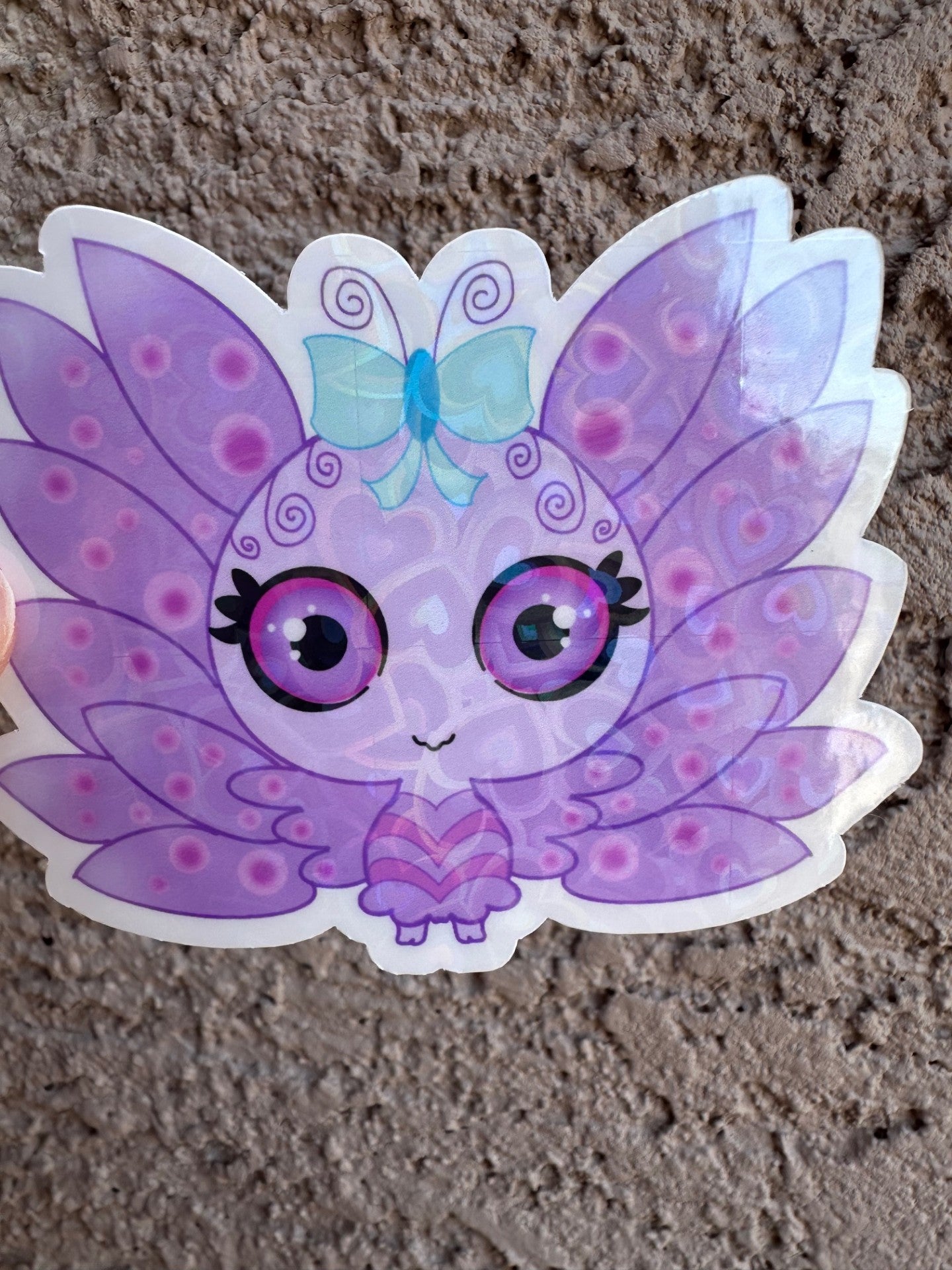 A vinyl heart holographic sticker cartoon butterfly character with purple wings and a blue bow. The character has purple and black eyes with light purple circle patterns on the wings. The background is a textured brown background.