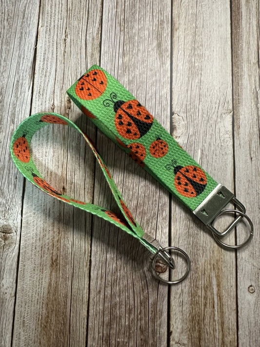 A fabric wristlet keychain featuring a green background patterned with black and red ladybugs, shown on a light wooden surface.