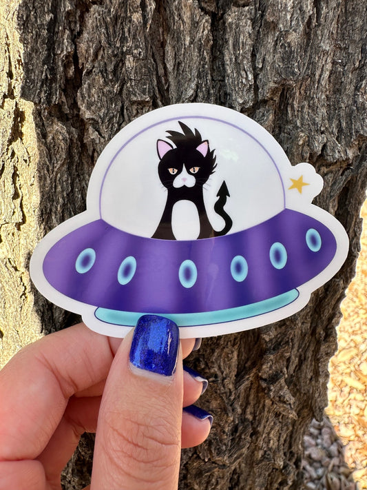 Sticker of a cat inside a UFO that is purple and turquoise in color, held by a hand against a tree background.