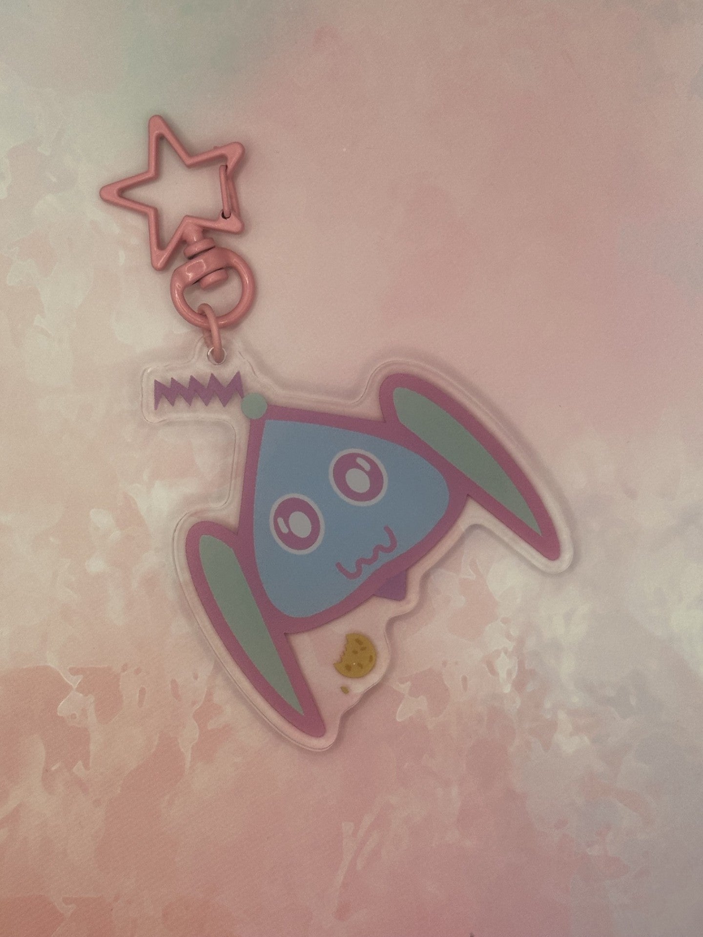 Fictional character keychain that is light blue and turquoise that has big and white eyes, with a purple antenna on head and has a cookie next to it. The keychain has a pink star key ring. The acrylic keychain is sitting on top of a pink textured background.