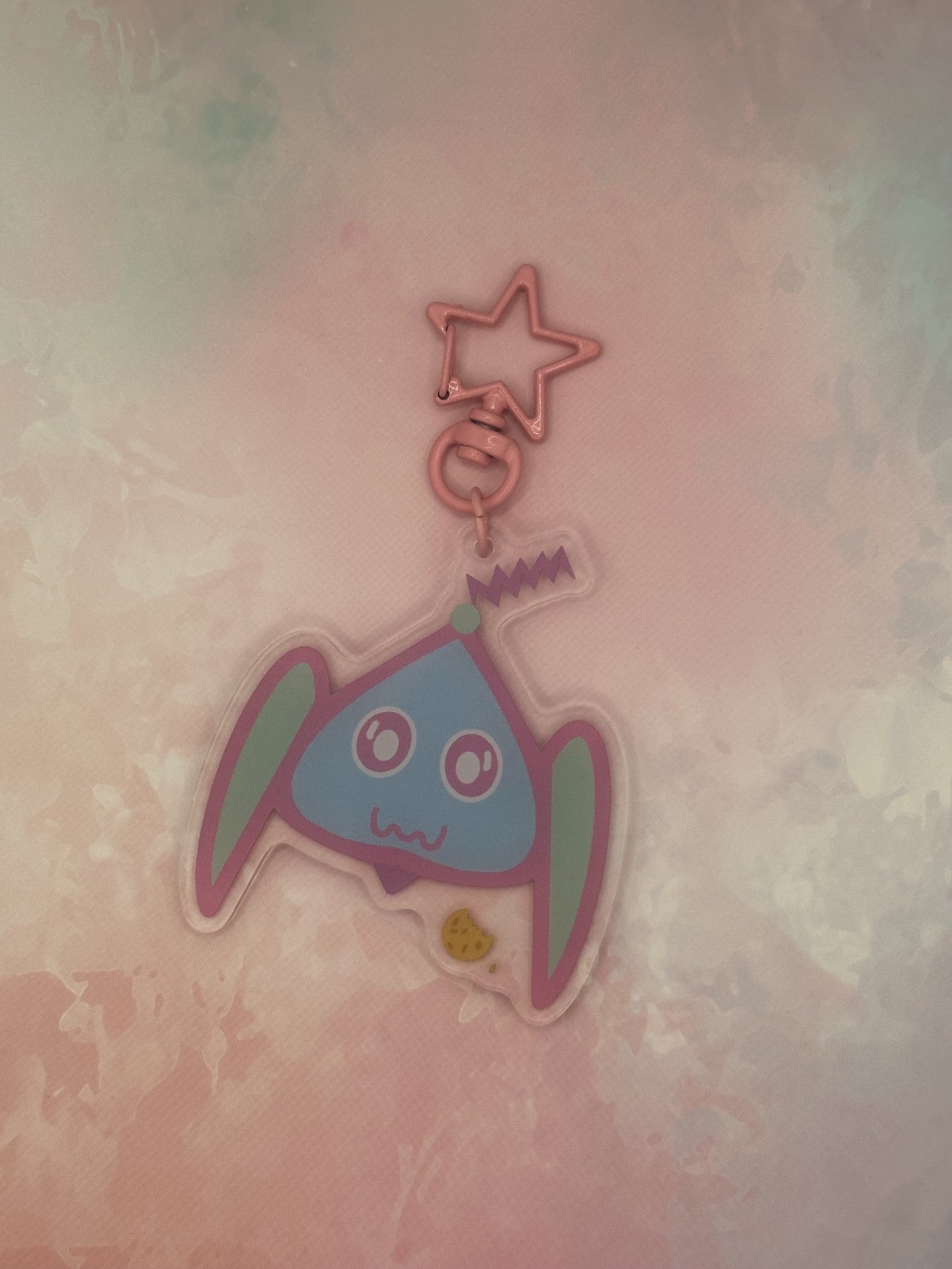 Fictional character keychain that is light blue and turquoise that has big and white eyes, with a purple antenna on head and has a cookie next to it. The keychain has a pink star key ring. The acrylic keychain is sitting on top of a pink textured background.