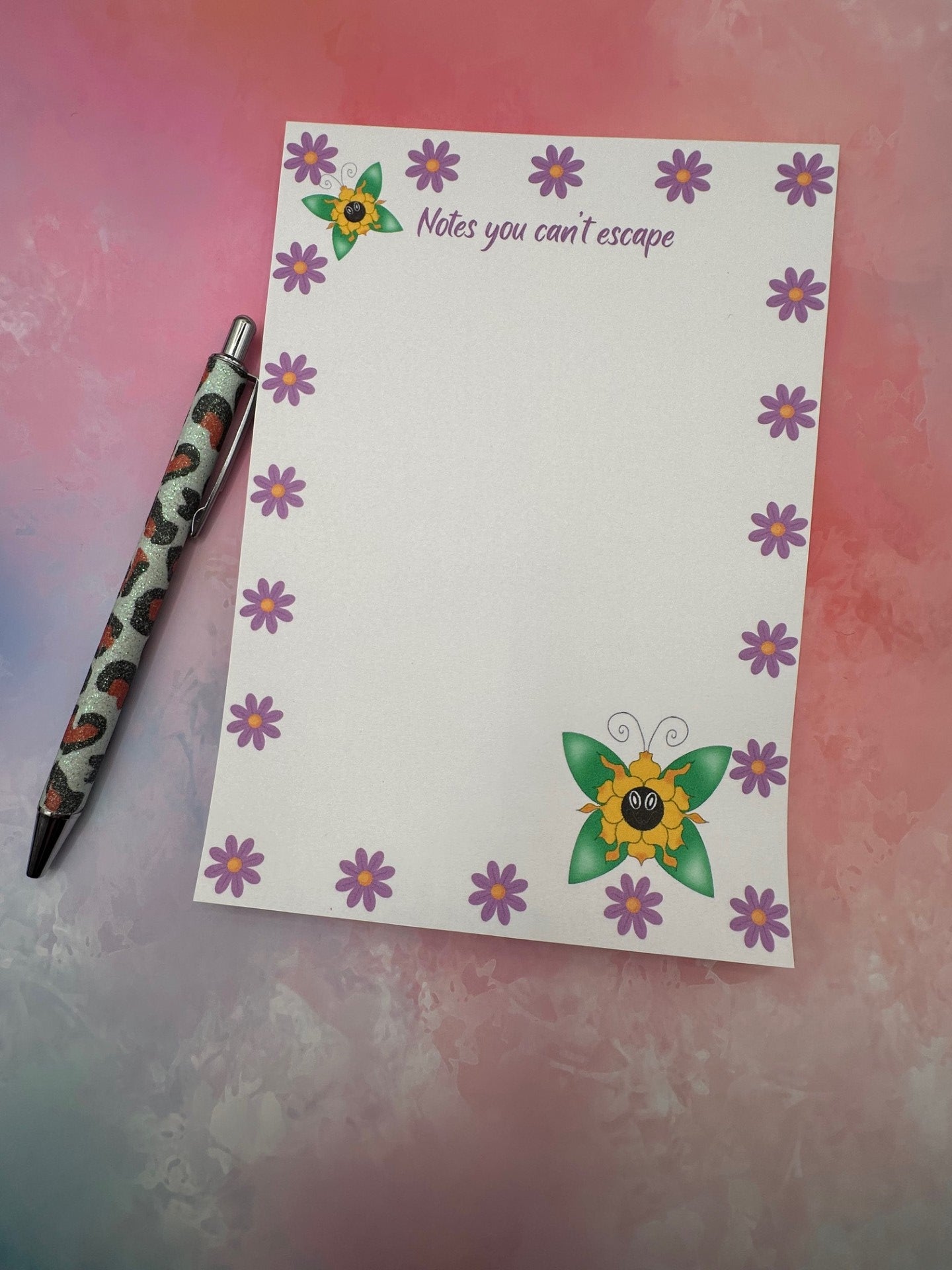 A notepad with fictional character that is a yellow flowery shape and has green wings coming from it. The notepad all around has purple flowers with a heading of "notes you can't escape". The notepad is sitting on top of a pink background with a pen.