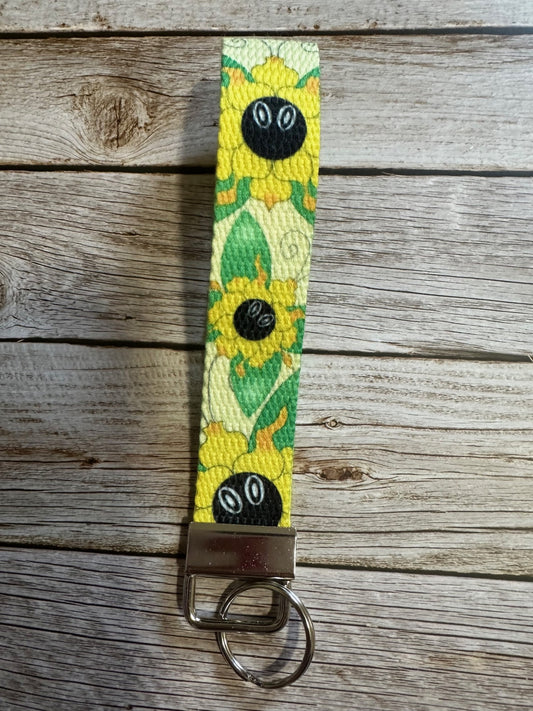 Wristlet keychain with sunflower design that also has butterfly wings coming from it. The wrislet is sitting on a wooden surface. 