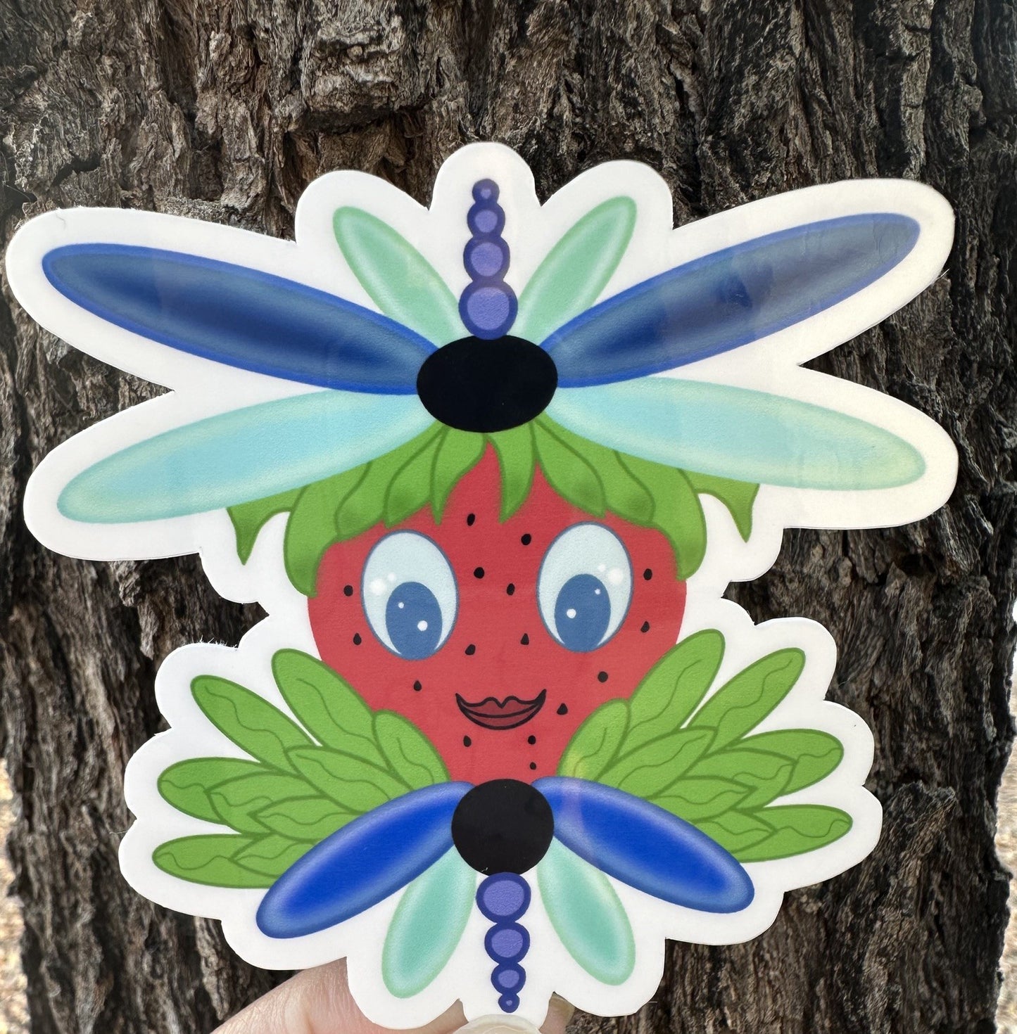 Sticker of a strawberry with dragonfly wings that is blue and light turquoise in color. The character has green leaves coming out of it. The sticker background is a tree trunk. 