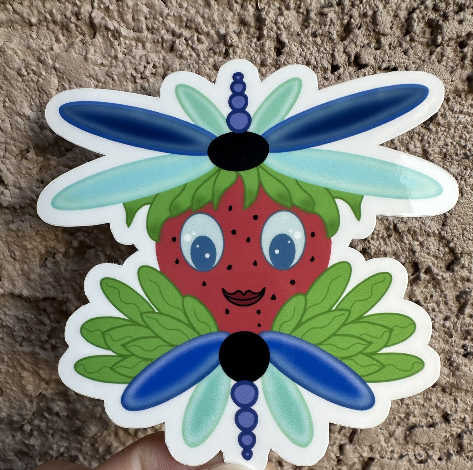Sticker of a strawberry with dragonfly wings that is blue and light turquoise in color. The character has green leaves coming out of it. The sticker background is a textured wall.