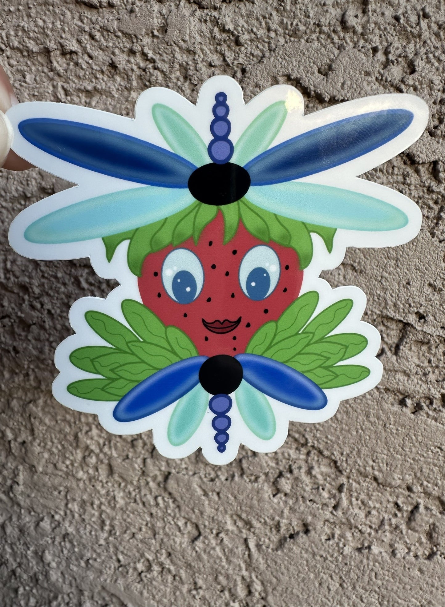 Sticker of a strawberry with dragonfly wings that is blue and light turquoise in color. The character has green leaves coming out of it. The sticker background is a textured wall.