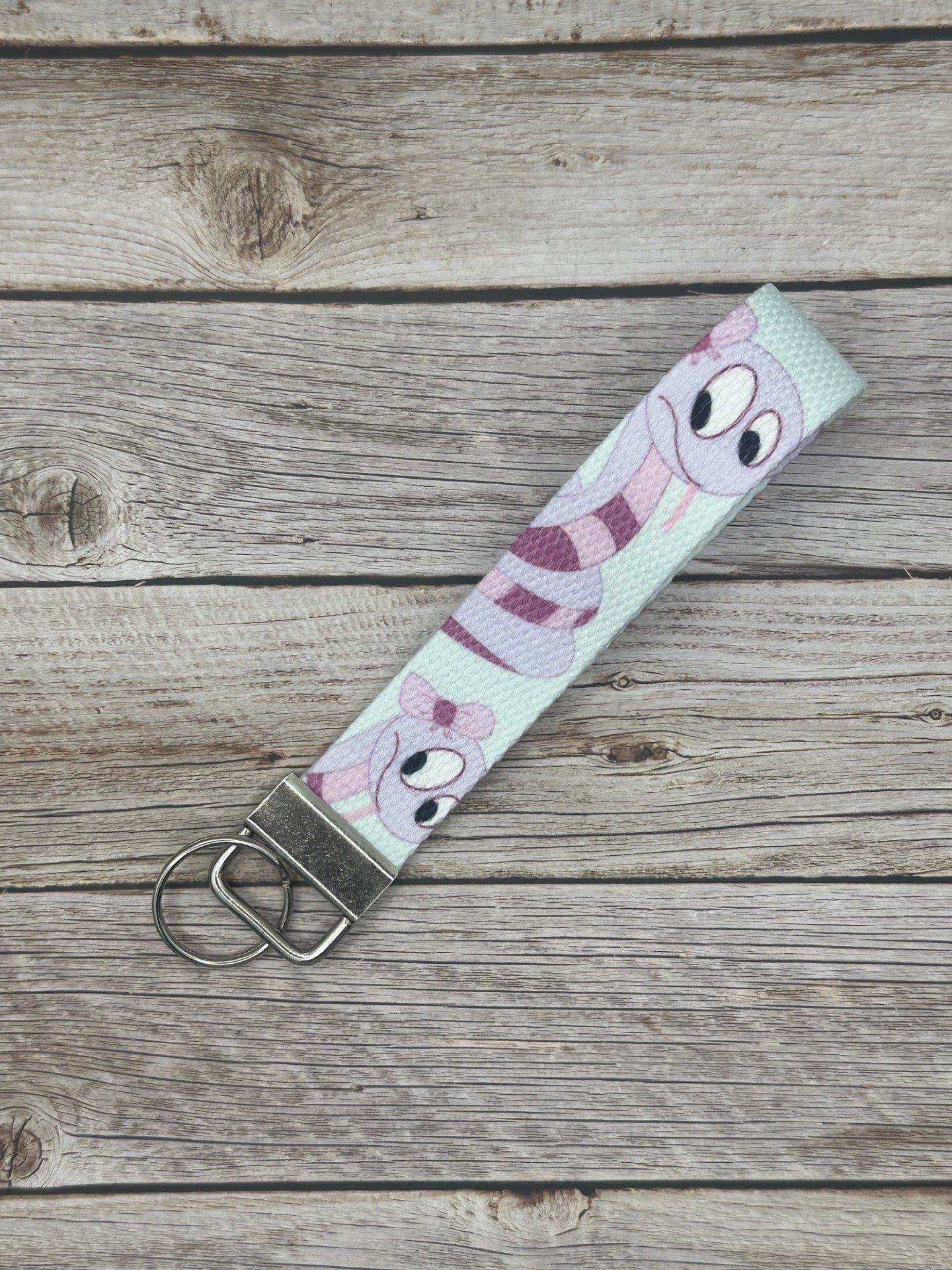 Keychain with a purple cartoon character that is a snake and has dark and light purple colors, with a bow on its head. The wristlet is on a wooden surface.