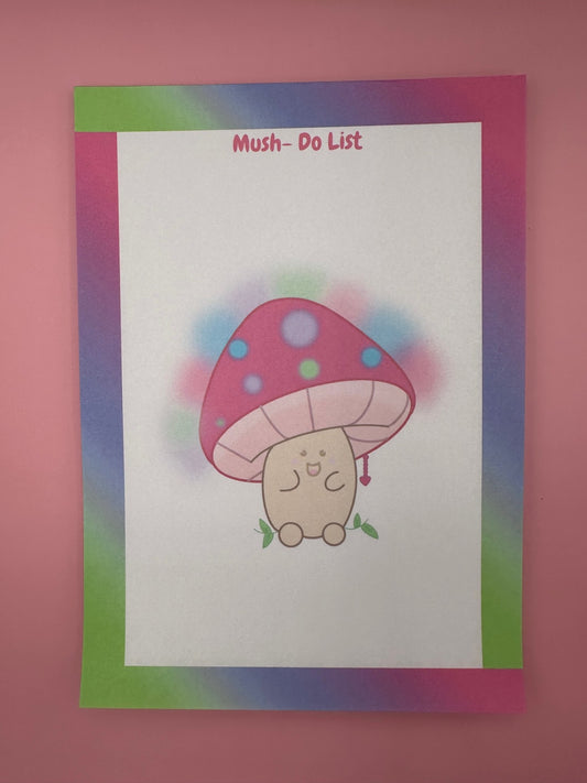 A notepad that has a character that is shaped like a mushroom with a smile face. The mushroom has different bright colors with a heart chain coming from it. The notepad has bright colors all around the notepad edges with the heading on the notepad "Mush-Do List". The notepad is sitting on top of a pink background with a pen. 
