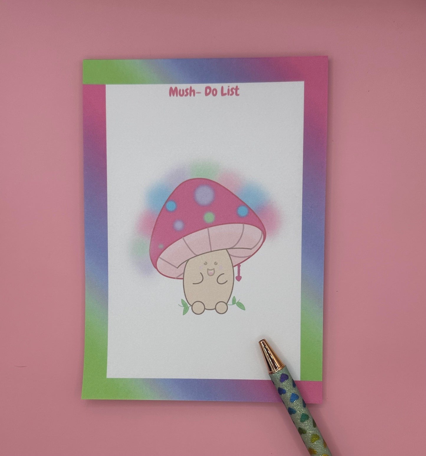 A notepad that has a character that is shaped like a mushroom with a smile face. The mushroom has different bright colors with a heart chain coming from it. The notepad has bright colors all around the notepad edges with the heading on the notepad "Mush-Do List". The notepad is sitting on top of a pink background with a pen. 