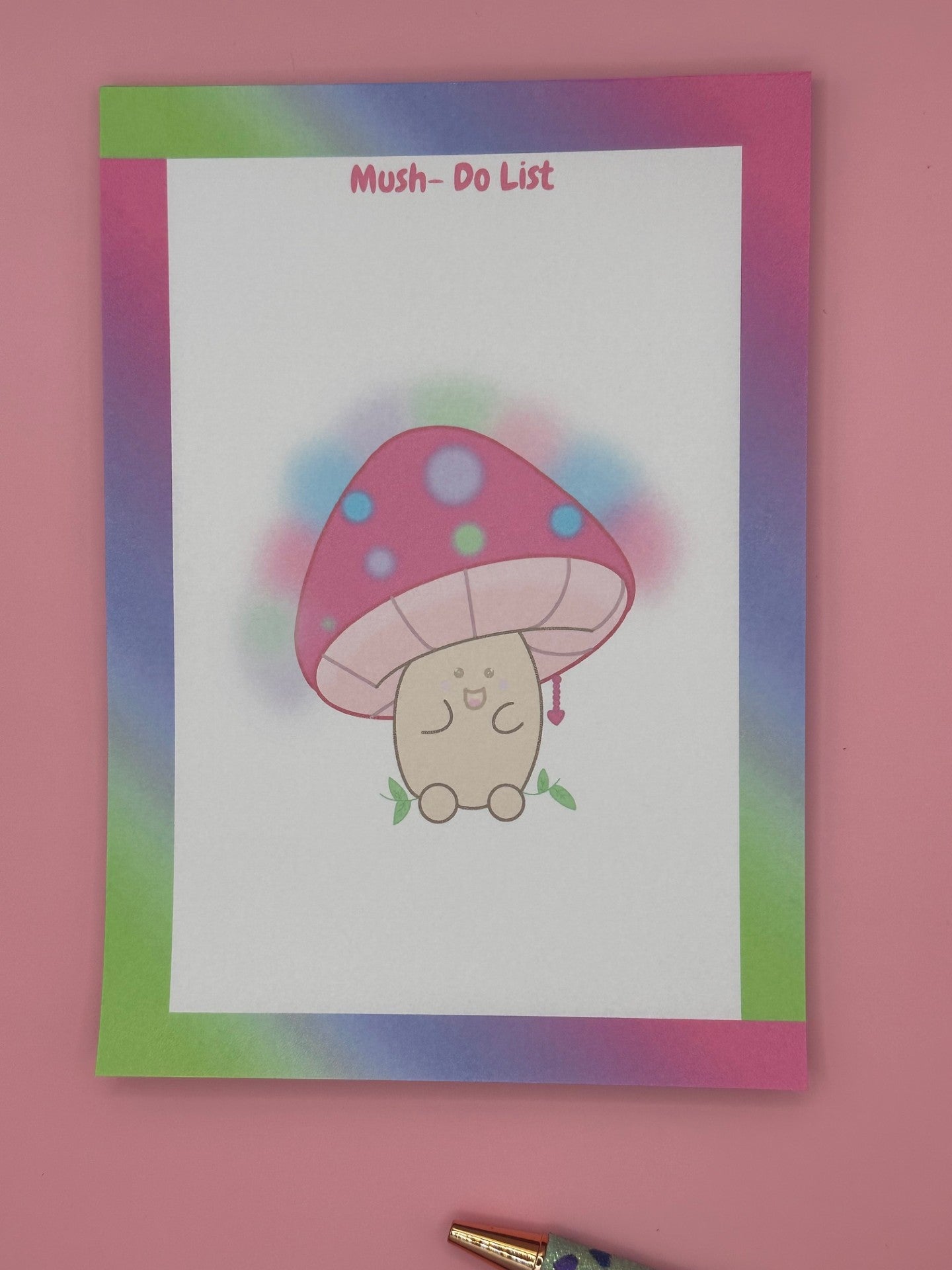 A notepad that has a character that is shaped like a mushroom with a smile face. The mushroom has different bright colors with a heart chain coming from it. The notepad has bright colors all around the notepad edges with the heading on the notepad "Mush-Do List". The notepad is sitting on top of a pink background with a pen. 