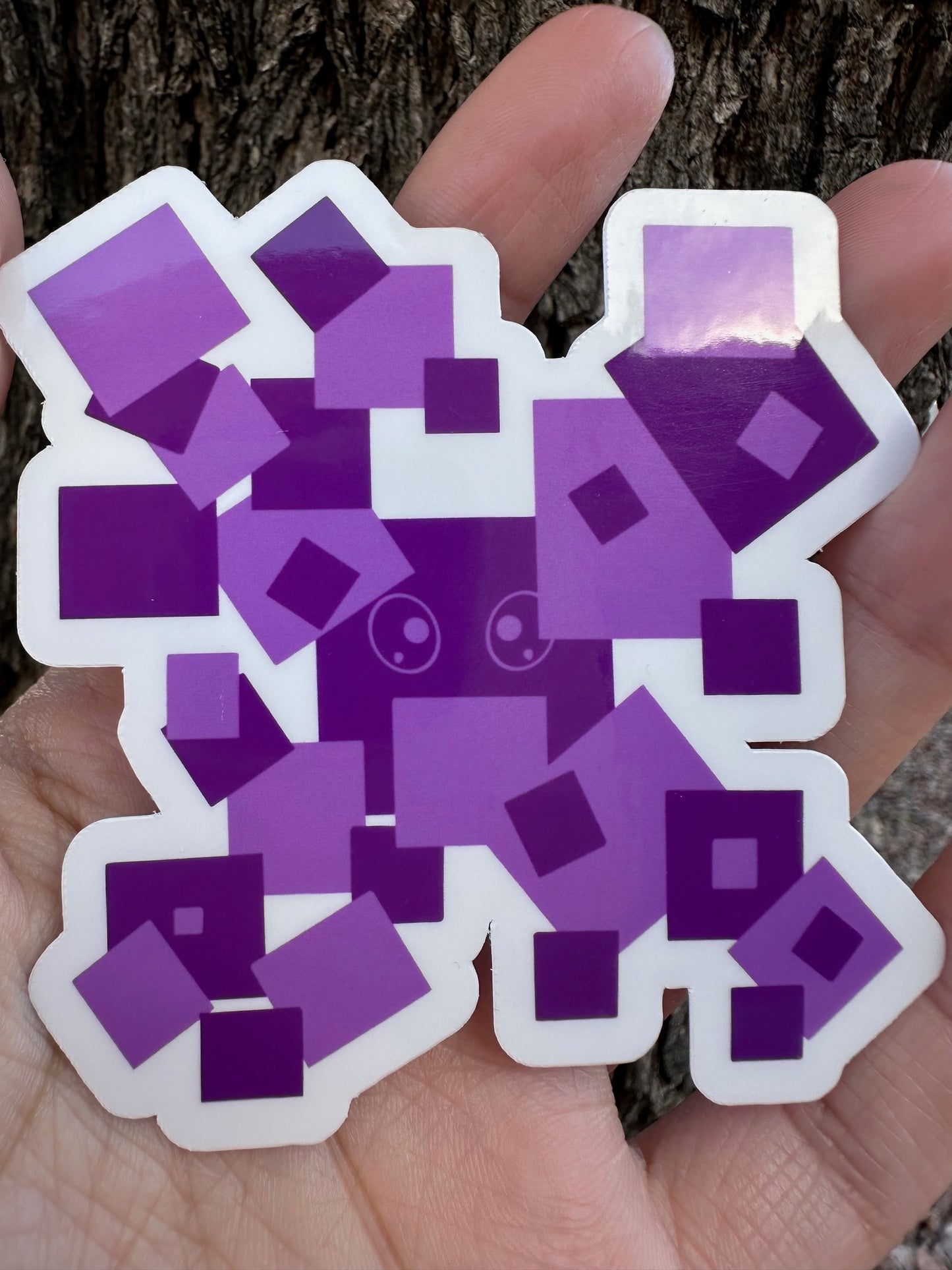A sticker featuring a pixelated character made of various overlapping purple squares, with a small cartoon face visible in the center.