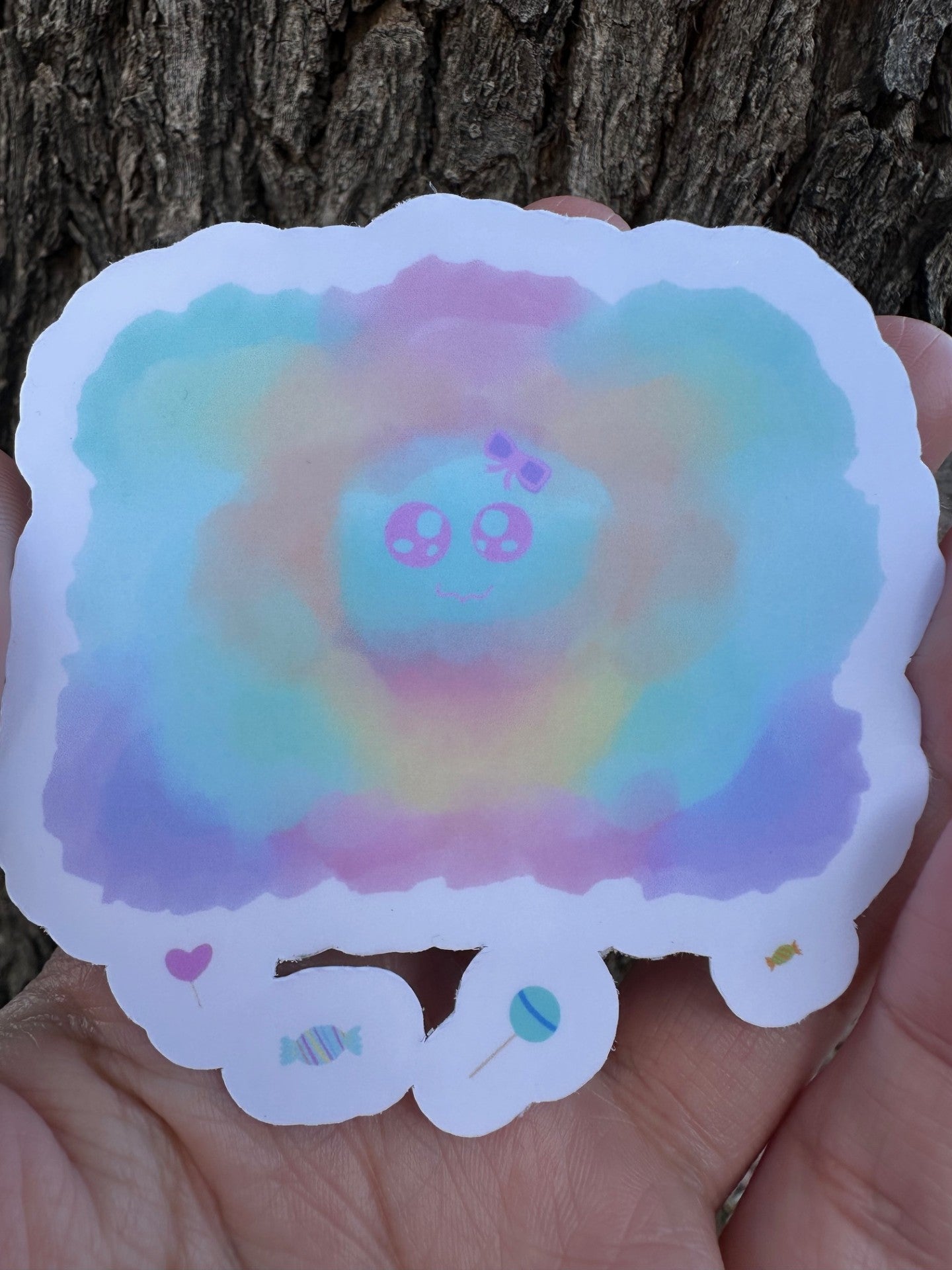 A sticker of a colorful, watercolor-style cloud shape in pastel blue, pink, yellow, and purple, featuring a cartoon face in the center and small candy illustrations along the bottom edge.