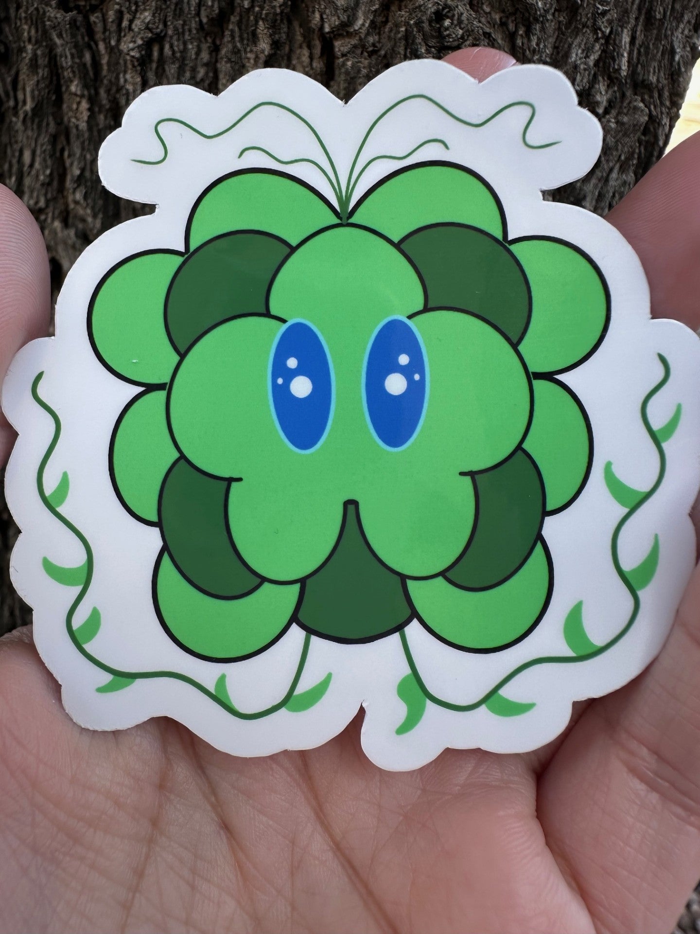 Green cartoonist character with blue eyes and small decorative vine illustrations held against a tree bark background.