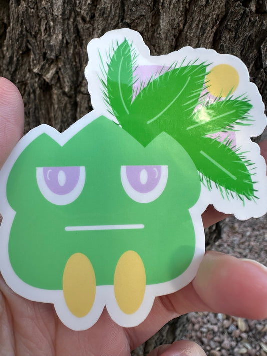 Cartoonish green creature sticker held by a hand against a tree bark background.