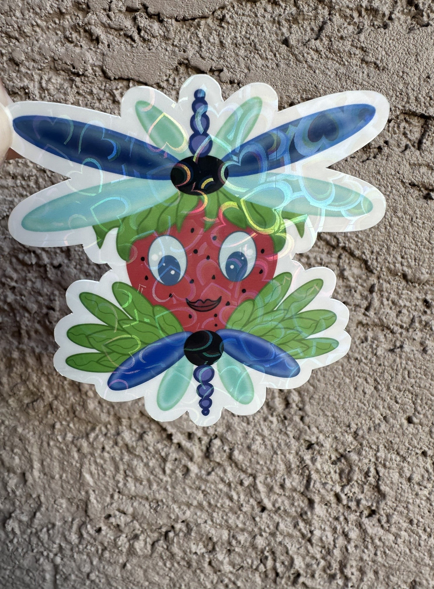 Sticker of a strawberry with dragonfly wings that is blue and light turquoise in color. The character has green leaves coming out of it. The sticker background is a textured wall.