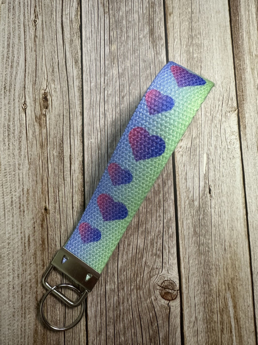 A wristlet keychain featuring a blue-to-green gradient background and a pattern of small, multi-color hearts and stars laid on a light wooden surface.