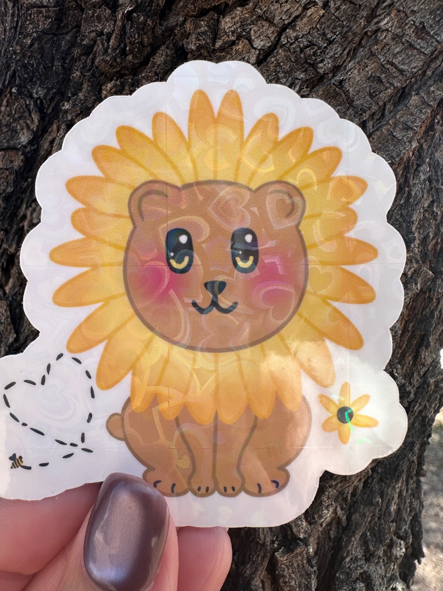Sticker with a brown bear that has rosy cheeks, with black and yellow eyes and a black mouth. The character has a sunflower surrounding the head. Besides the character is a bumble bee with a heart and a sunflower. The sticker has a white outline and the background is a tree bark. 