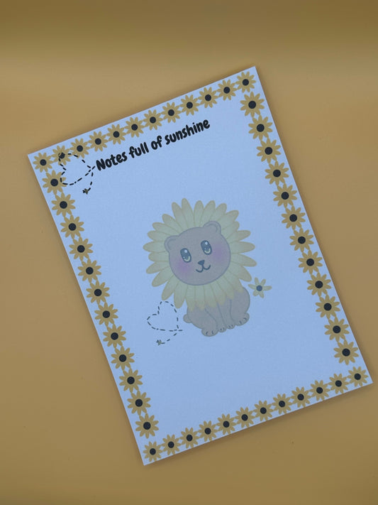 A notepad with a fictional character that looks like a brown bear with a yellow flower around its head. The character has yellow and black eyes with a black mouth. The character has a yellow flower next to it and a bumble bee with a black heart. The notepad edges all around four corners have yellow sunflowers and the same bumble bee heart design on top. The notepad heading is " Notes full of sunshine". The notepad is sitting on top of a yellow background. 