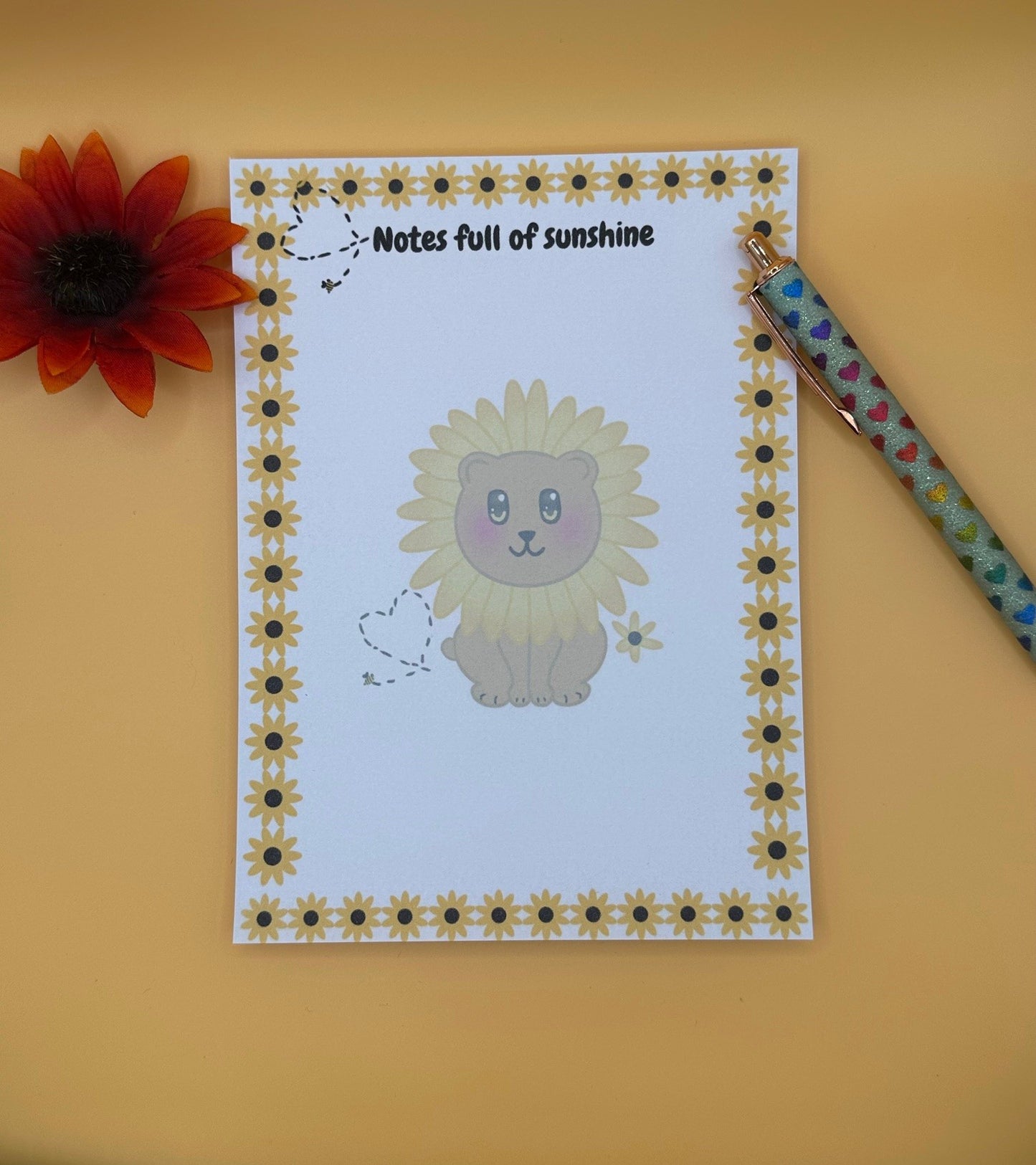 A notepad with a fictional character that looks like a brown bear with a yellow flower around its head. The character has yellow and black eyes with a black mouth. The character has a yellow flower next to it and a bumble bee with a black heart. The notepad edges all around four corners have yellow sunflowers and the same bumble bee heart design on top. The notepad heading is " Notes full of sunshine". The notepad is sitting on top of a yellow background with a pen and flower. 