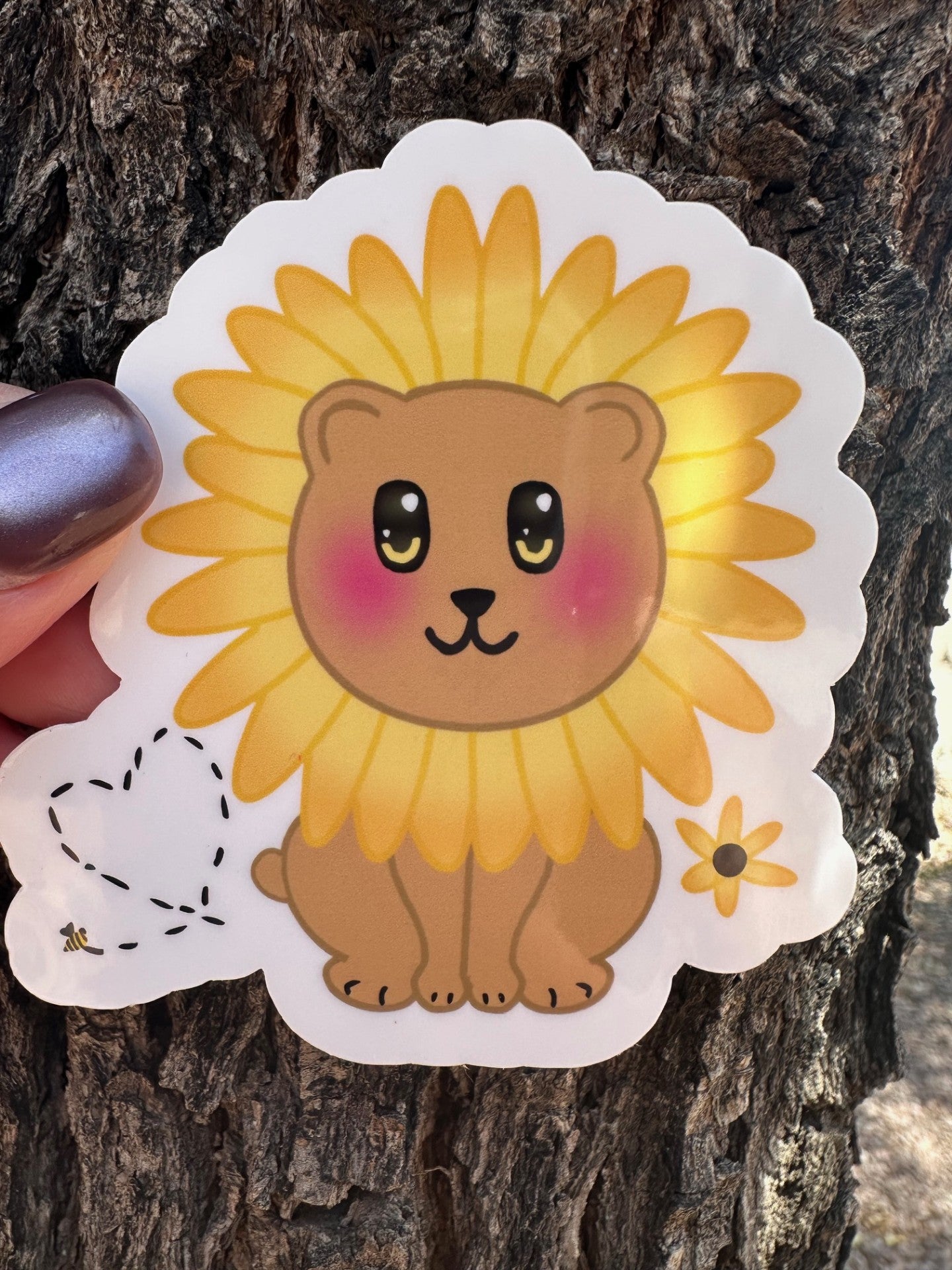 Sticker with a brown bear that has rosy cheeks, with black and yellow eyes and a black mouth. The character has a sunflower surrounding the head. Besides the character is a bumble bee with a heart and a sunflower. The sticker has a white outline and the background is a tree bark. 
