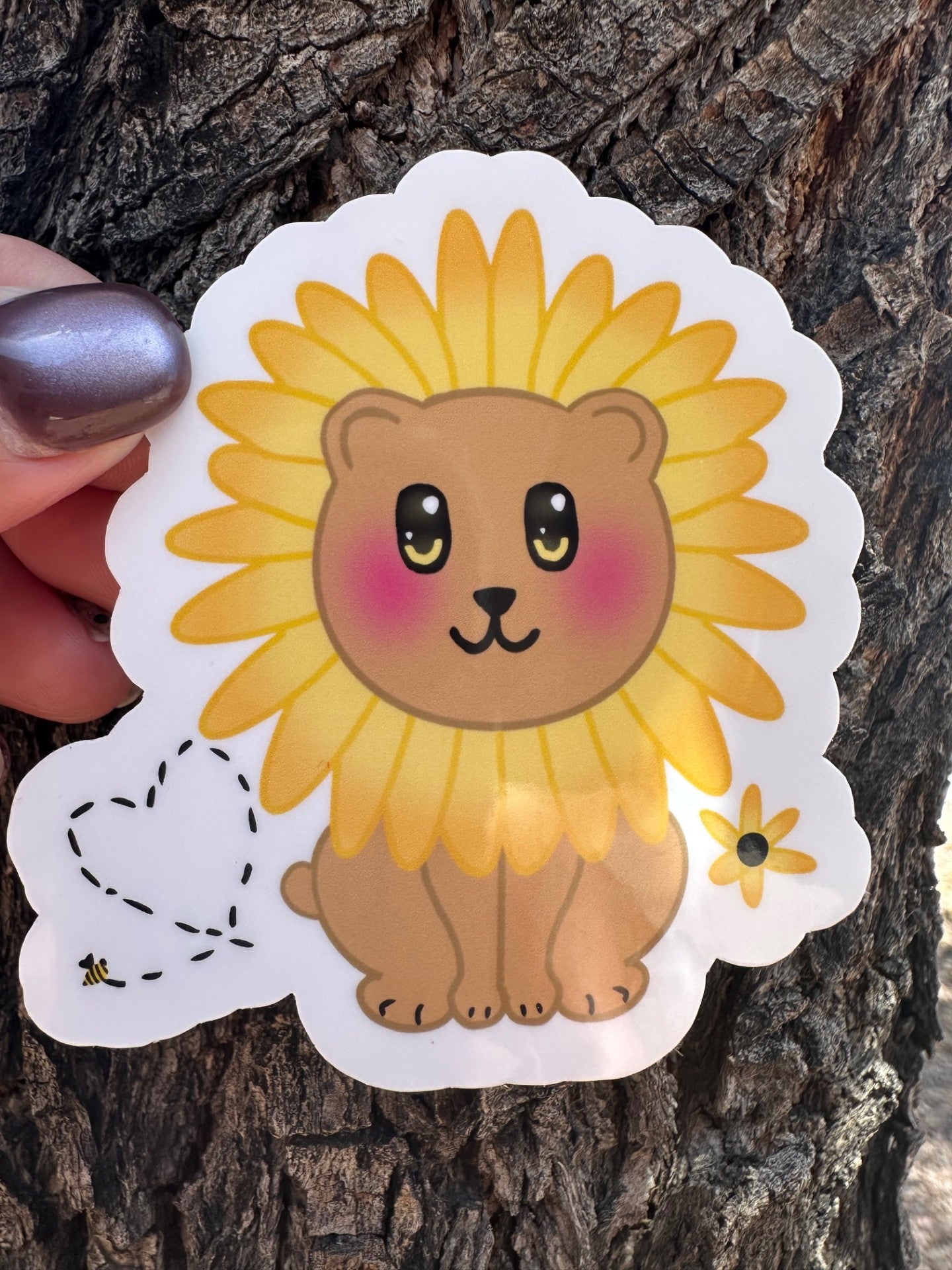 Sticker with a brown bear that has rosy cheeks, with black and yellow eyes and a black mouth. The character has a sunflower surrounding the head. Besides the character is a bumble bee with a heart and a sunflower. The sticker has a white outline and the background is a tree bark. 