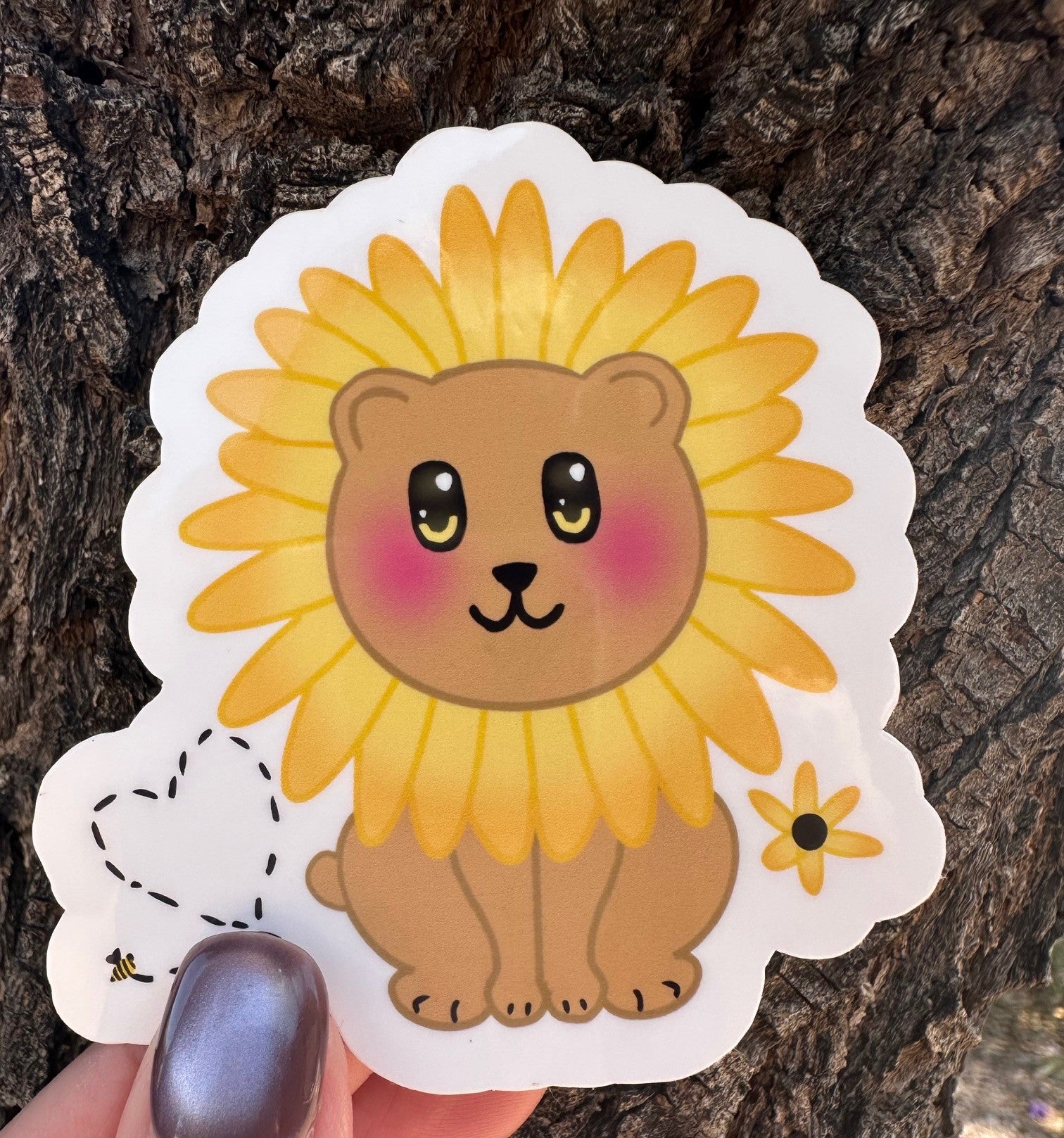 Sticker with a brown bear that has rosy cheeks, with black and yellow eyes and a black mouth. The character has a sunflower surrounding the head. Besides the character is a bumble bee with a heart and a sunflower. The sticker has a white outline and the background is a tree bark. 