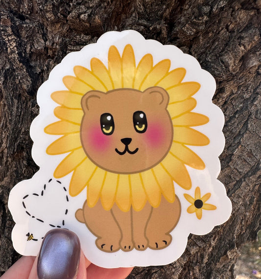 Sticker with a brown bear that has rosy cheeks, with black and yellow eyes and a black mouth. The character has a sunflower surrounding the head. Besides the character is a bumble bee with a heart and a sunflower. The sticker has a white outline and the background is a tree bark. 