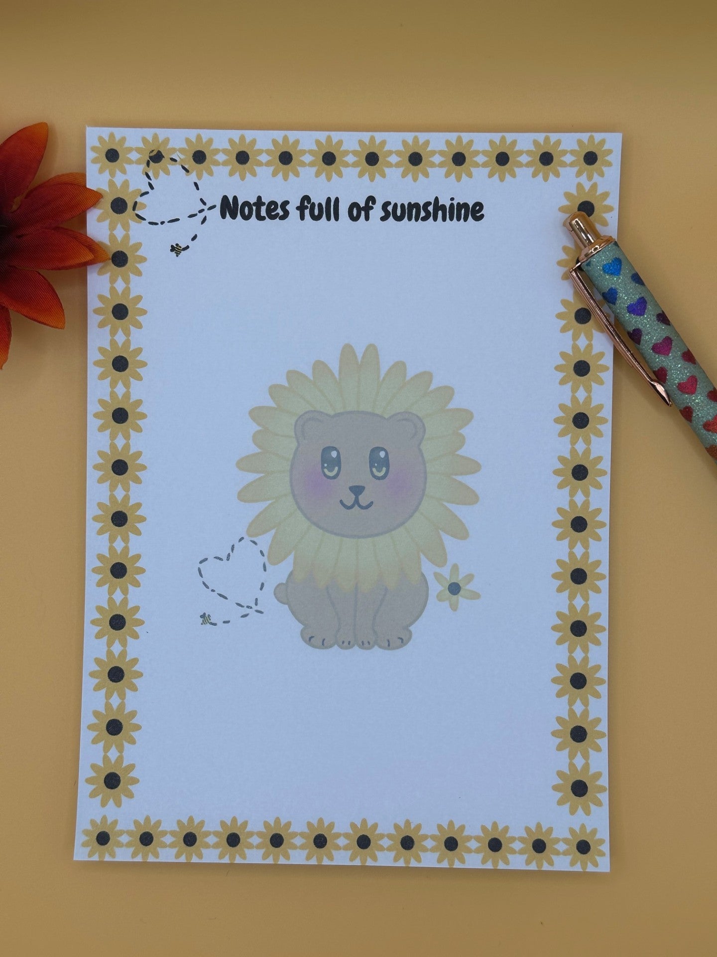A notepad with a fictional character that looks like a brown bear with a yellow flower around its head. The character has yellow and black eyes with a black mouth. The character has a yellow flower next to it and a bumble bee with a black heart. The notepad edges all around four corners have yellow sunflowers and the same bumble bee heart design on top. The notepad heading is " Notes full of sunshine". The notepad is sitting on top of a yellow background with a pen and flower. 
