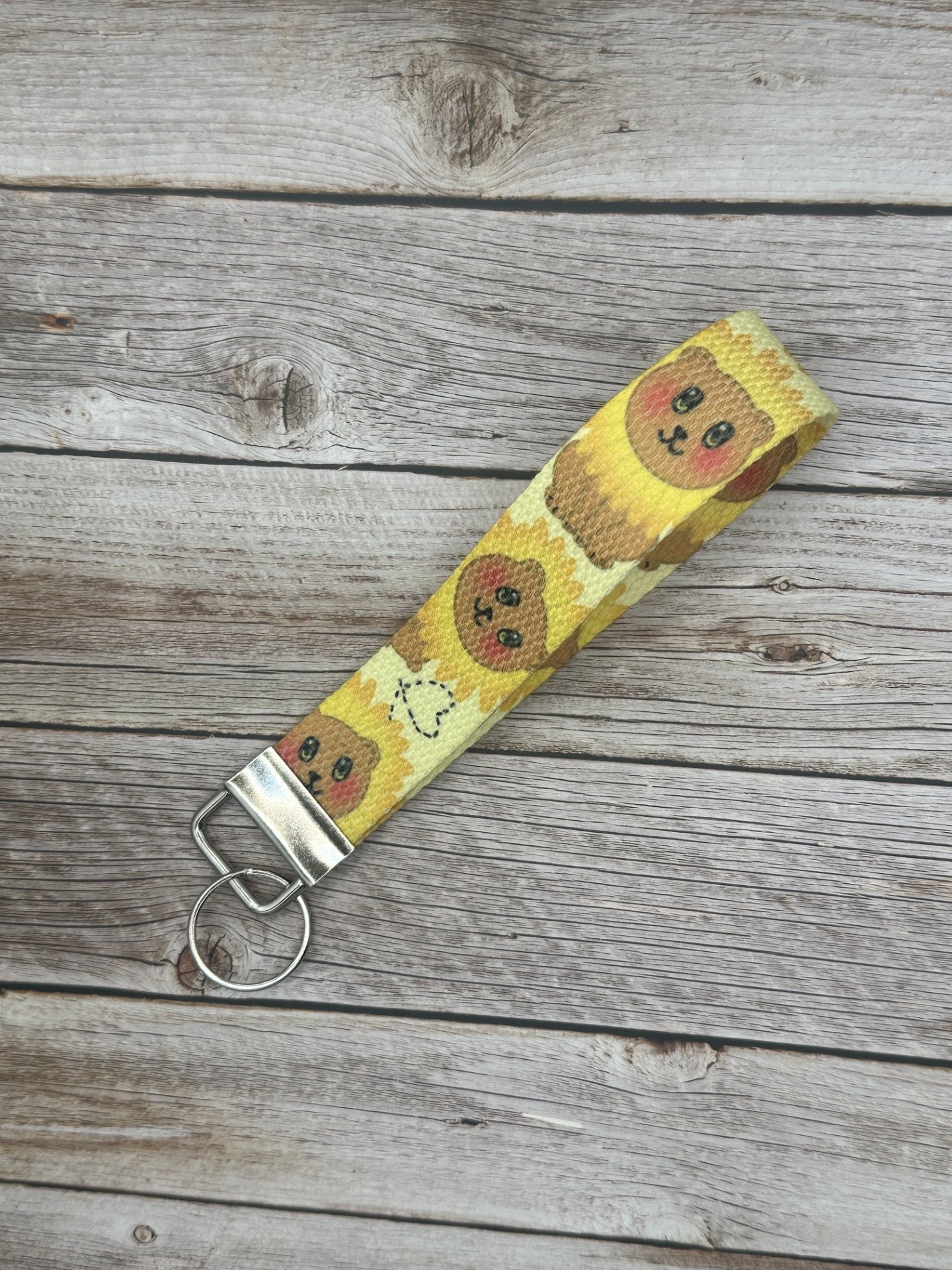 Wristlet keychain with a brown bear that has black eyes and rosy cheeks. The bear has a sunflower shape around its head, and a heart on the bottom of the wristlet. The keychain is on a wooden surface.