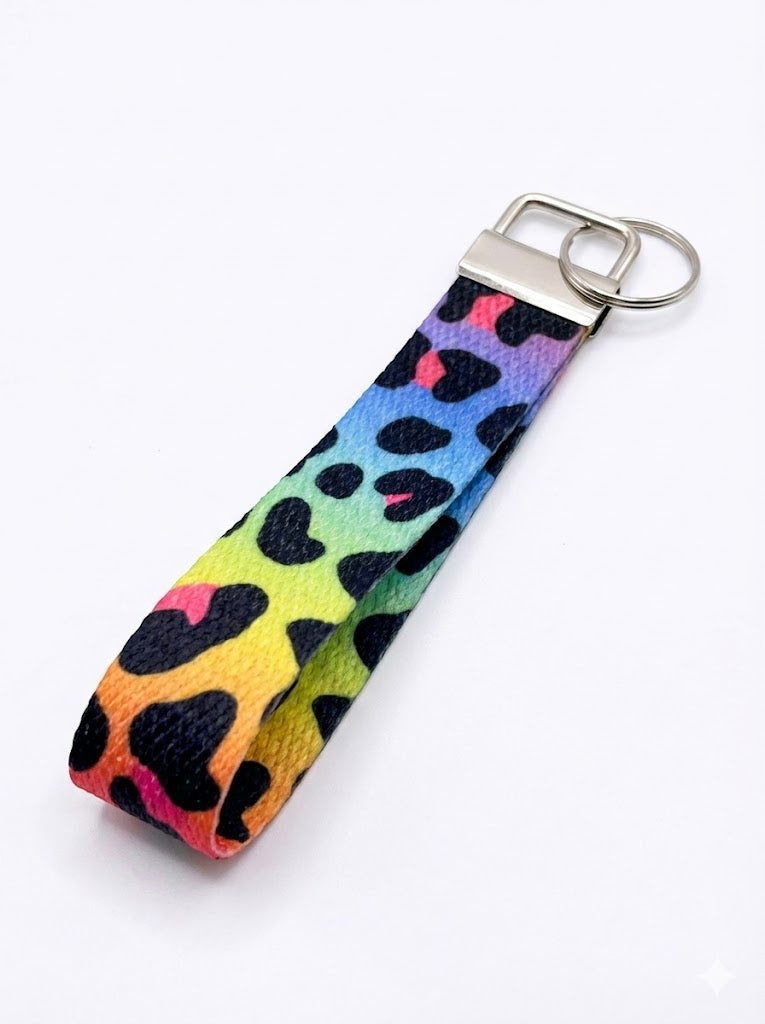 A fabric wristlet keychain with a rainbow gradient background in yellow, green, blue, and purple, featuring a black and pink leopard print pattern and a silver-toned metal key ring attachment.
