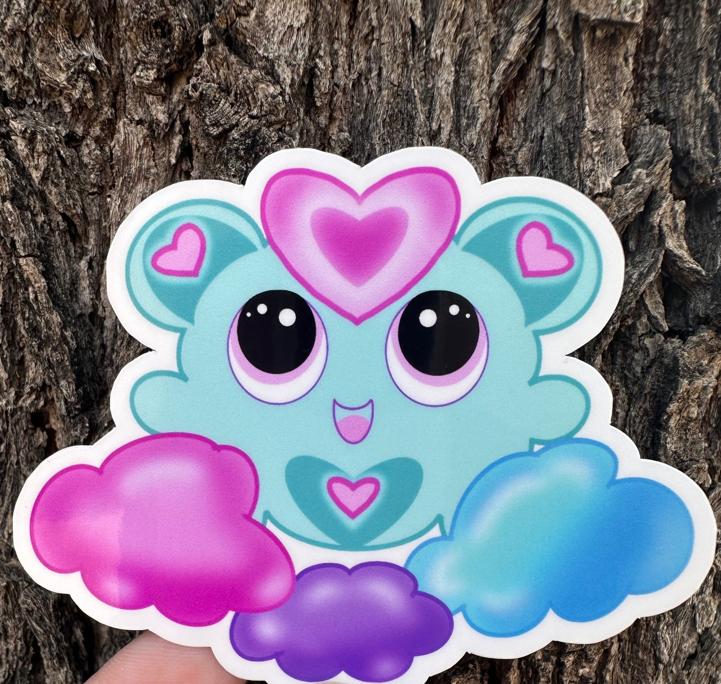 A fictional Cartoon character with heart-shaped ears that has different colors. The colors on the character are blue, pink, and purple pastel. The character is on a colorful pastel of the same color, and the background is a tree branch. 