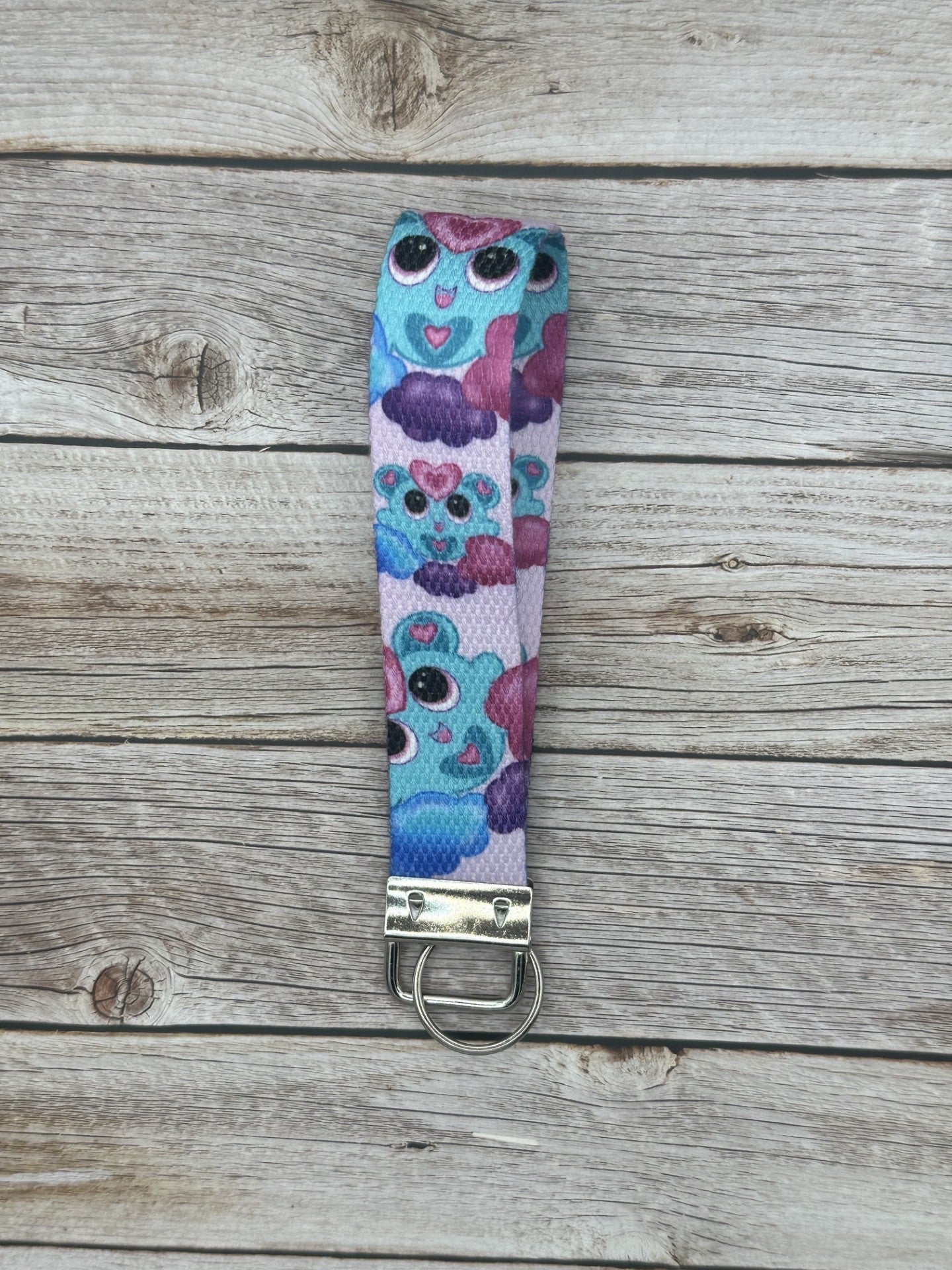 Unique fictional character that is blue and has pink and turquoise hearts on it. The character is also on a purple, pink and blue cloud and has black eyes with pink in them. The background color of the wristlet is light pink. The wristlet is sitting on top of a wooden background. 