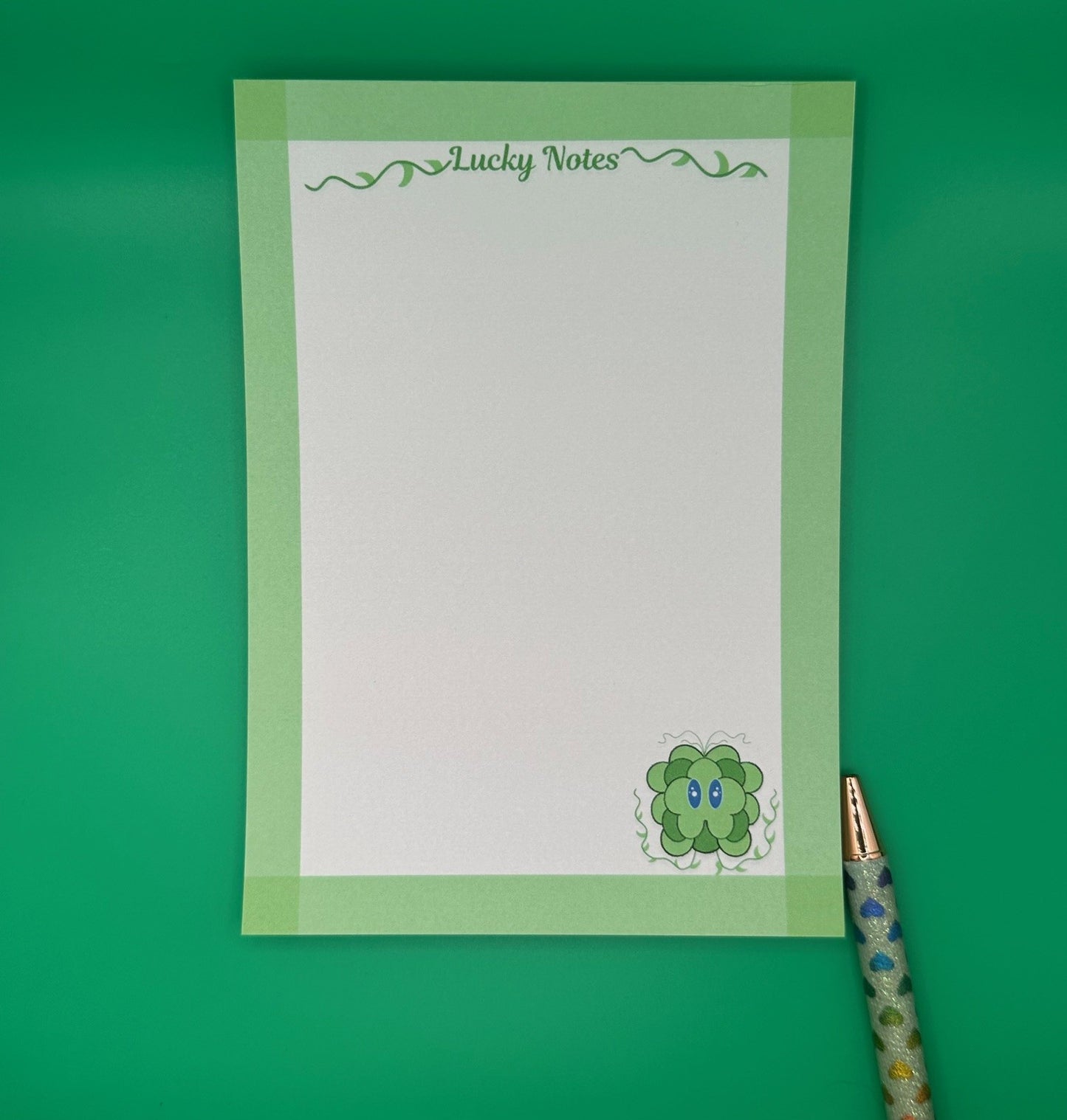 A notepad that has a green leave character at the bottom of the notepad. The character has blue eyes and green vines coming from it. The notepad on all four edges is green and has a heading with vines that states "Lucky notes". The notepad is sitting on top of a green background with a pen.