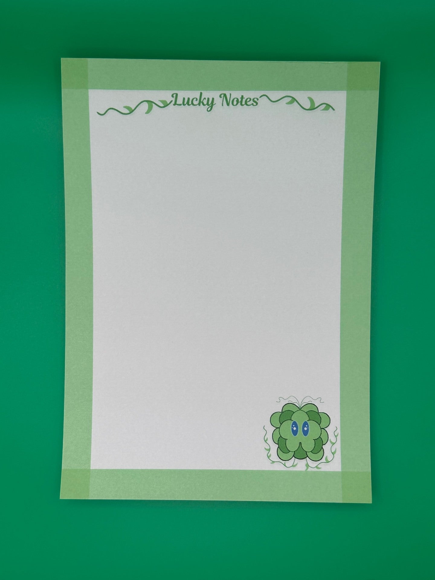A notepad that has a green leave character at the bottom of the notepad. The character has blue eyes and green vines coming from it. The notepad on all four edges is green and has a heading with vines that states "Lucky notes". The notepad is sitting on top of a green background.