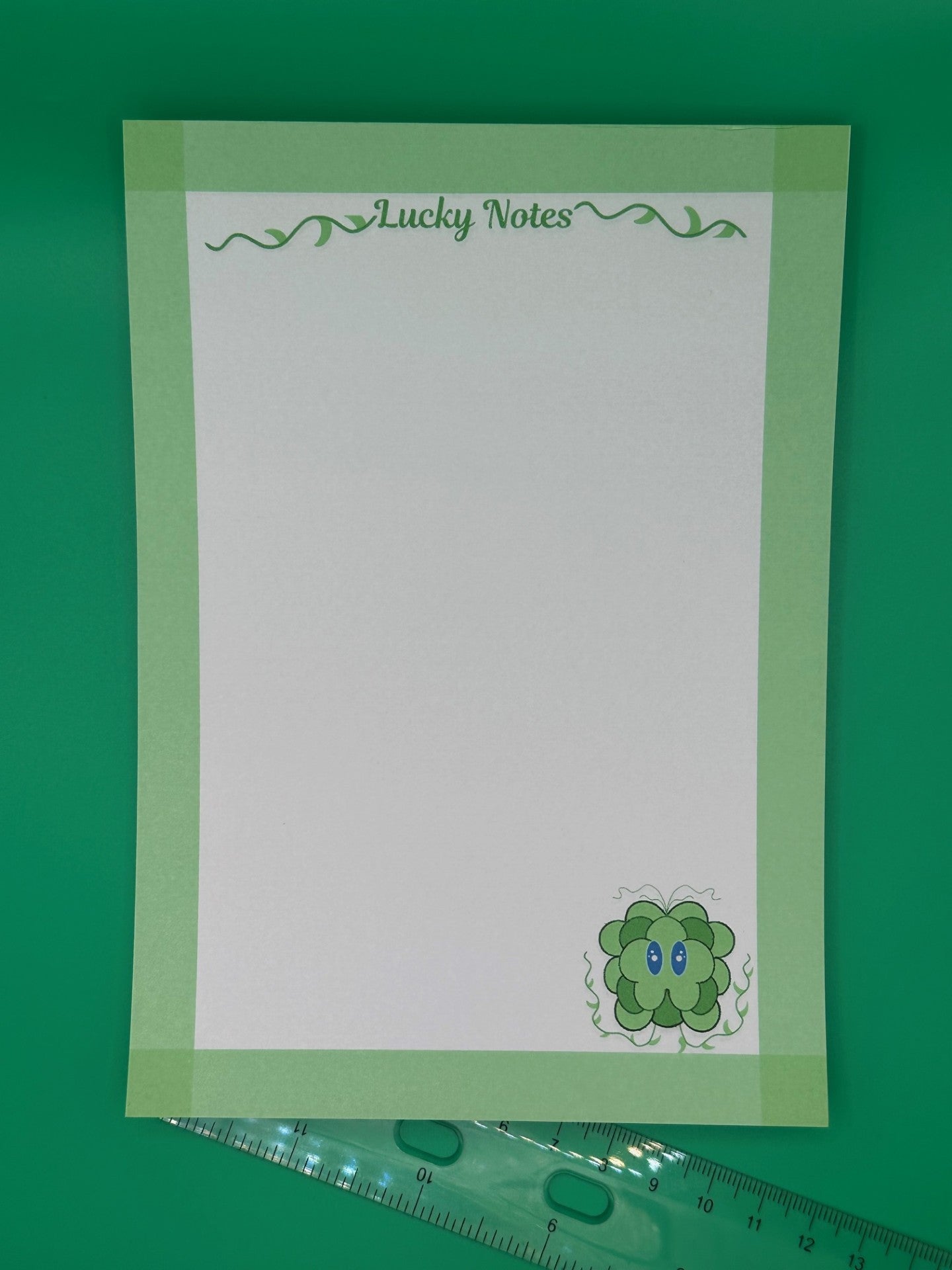 A notepad that has a green leave character at the bottom of the notepad. The character has blue eyes and green vines coming from it. The notepad on all four edges is green and has a heading with vines that states "Lucky notes". The notepad is sitting on top of a green background with a ruler. 