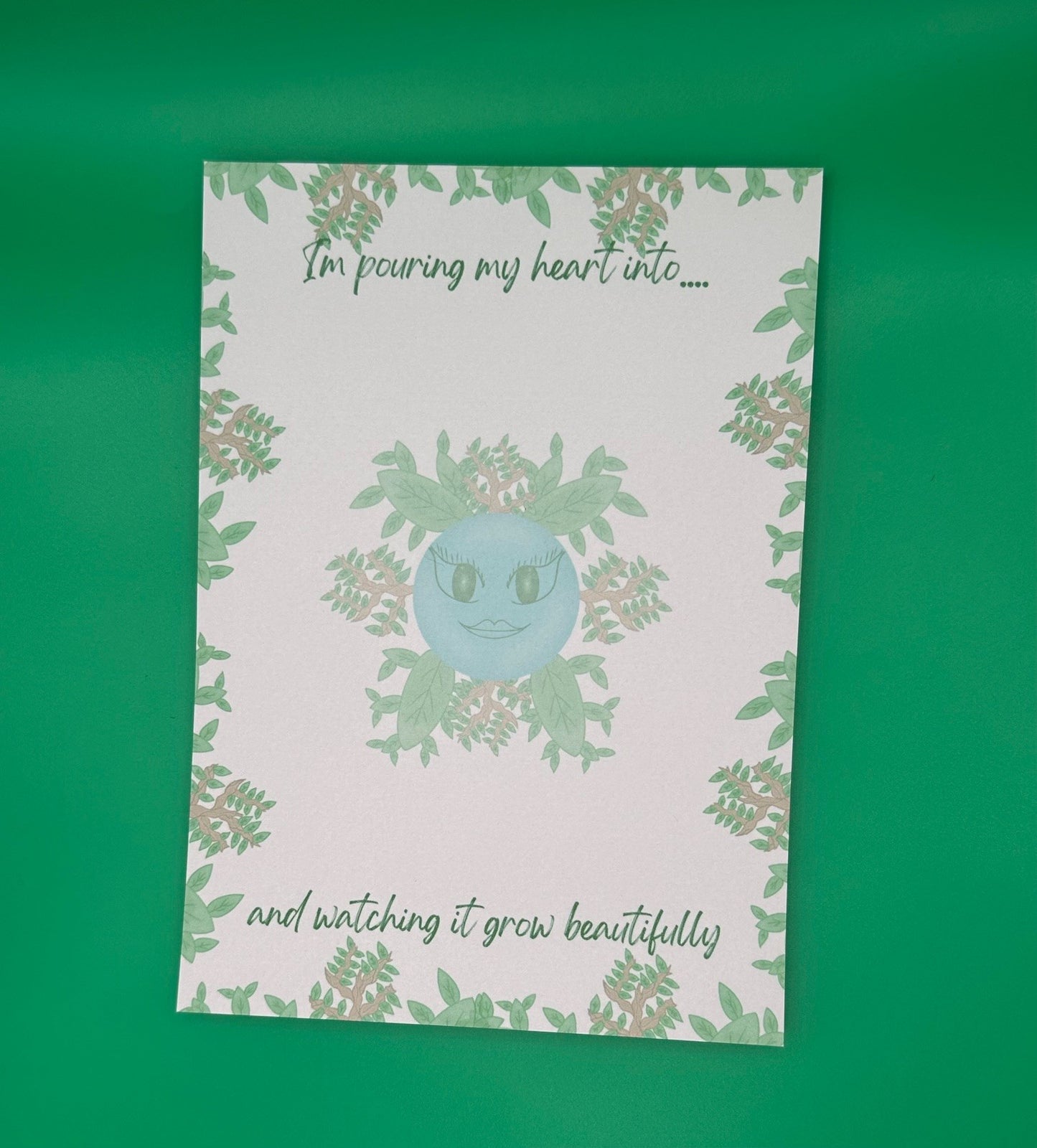 A notepad that has blue character with green eyes and lips. The character has leaves and branches coming out of it. The notepad has leaves and branches all around the edges. The notepad has a heading on top that states "I'm pouring my heart into...." and at the bottom states" and watching it grow beautifully". The notepad is sitting on top of a green background.