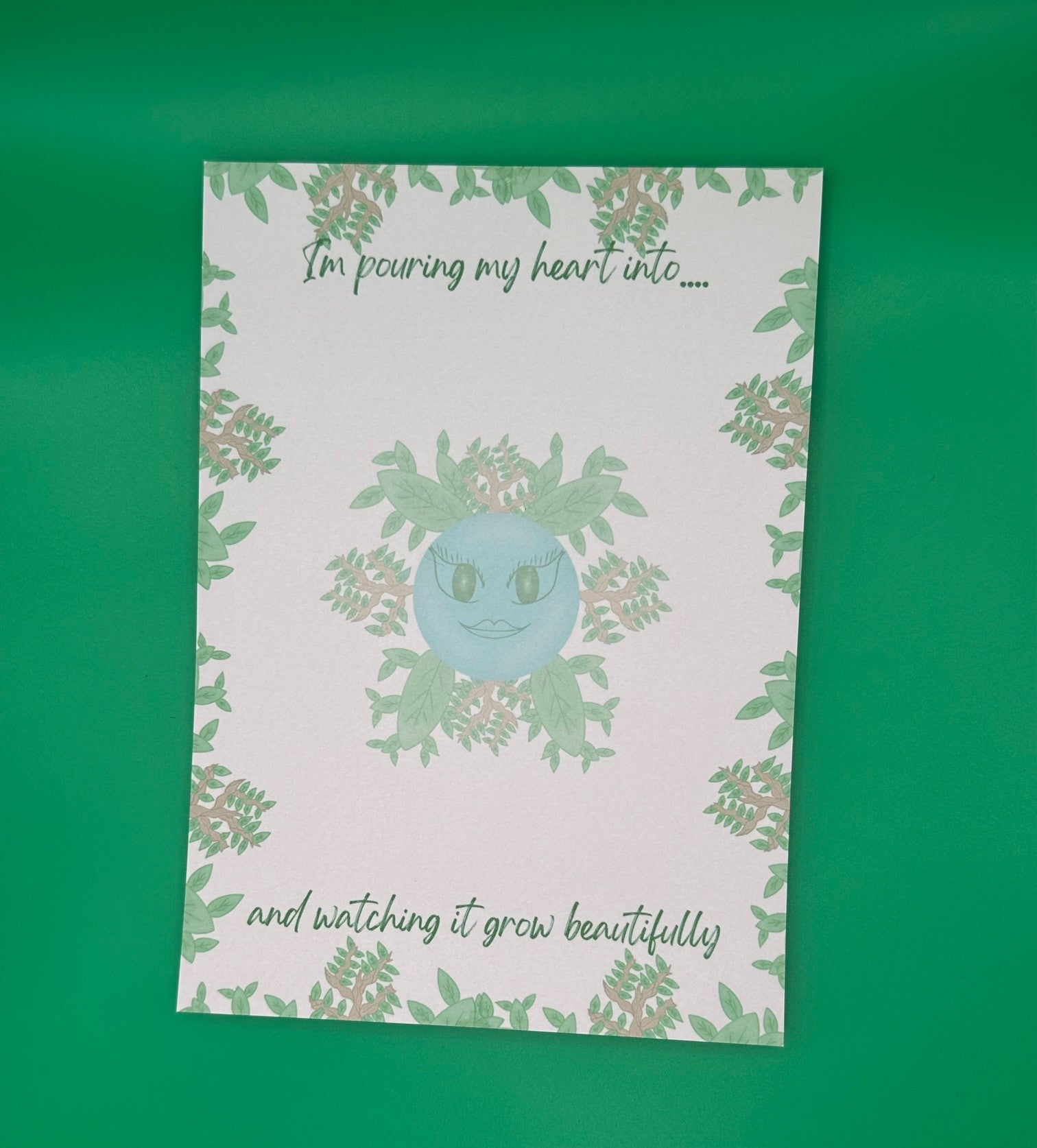 A notepad that has blue character with green eyes and lips. The character has leaves and branches coming out of it. The notepad has leaves and branches all around the edges. The notepad has a heading on top that states "I'm pouring my heart into...." and at the bottom states" and watching it grow beautifully". The notepad is sitting on top of a green background.