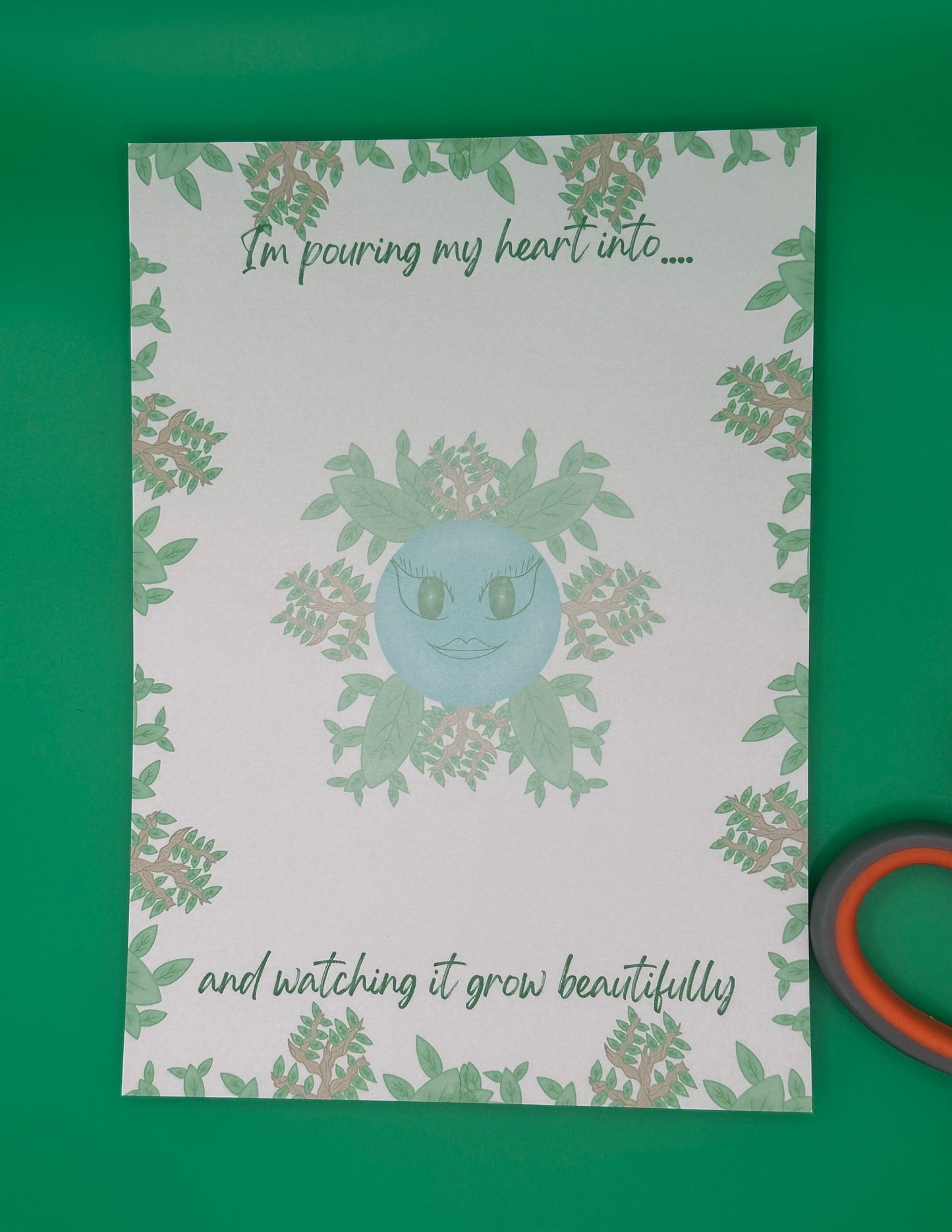 A notepad that has blue character with green eyes and lips. The character has leaves and branches coming out of it. The notepad has leaves and branches all around the edges. The notepad has a heading on top that states "I'm pouring my heart into...." and at the bottom states" and watching it grow beautifully". The notepad is sitting on top of a green background with scissors .