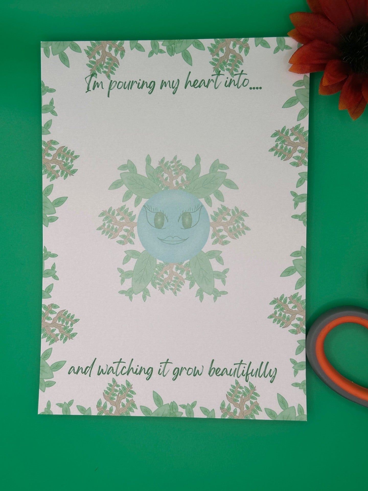 A notepad that has blue character with green eyes and lips. The character has leaves and branches coming out of it. The notepad has leaves and branches all around the edges. The notepad has a heading on top that states "I'm pouring my heart into...." and at the bottom states" and watching it grow beautifully". The notepad is sitting on top of a green background with scissors and a flower.