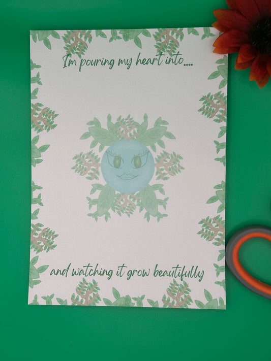 A notepad that has blue character with green eyes and lips. The character has leaves and branches coming out of it. The notepad has leaves and branches all around the edges. The notepad has a heading on top that states "I'm pouring my heart into...." and at the bottom states" and watching it grow beautifully". The notepad is sitting on top of a green background with scissors and a flower.